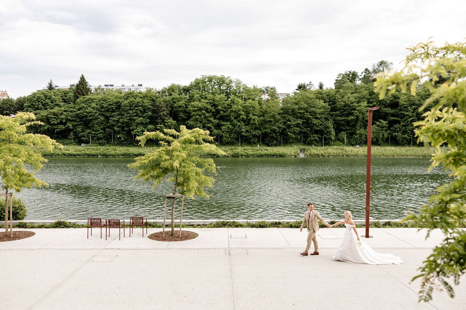 Bride and groom hold hands by a serene lakeside venue surrounded by lush green trees on their wedding day.