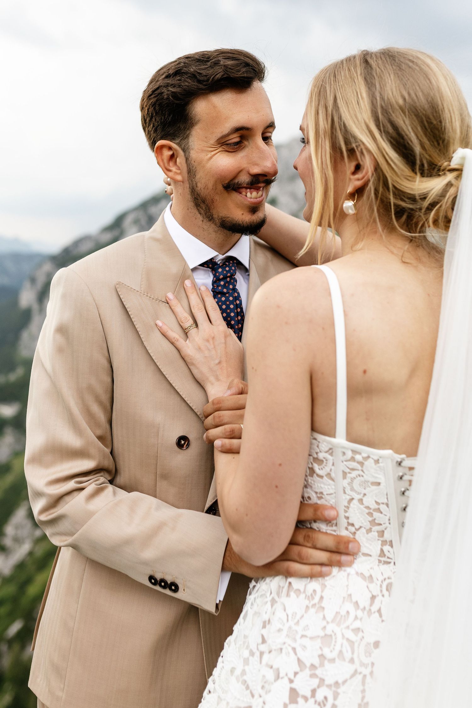 Smiling groom in beige suit embraces bride in lace wedding dress with mountain backdrop.