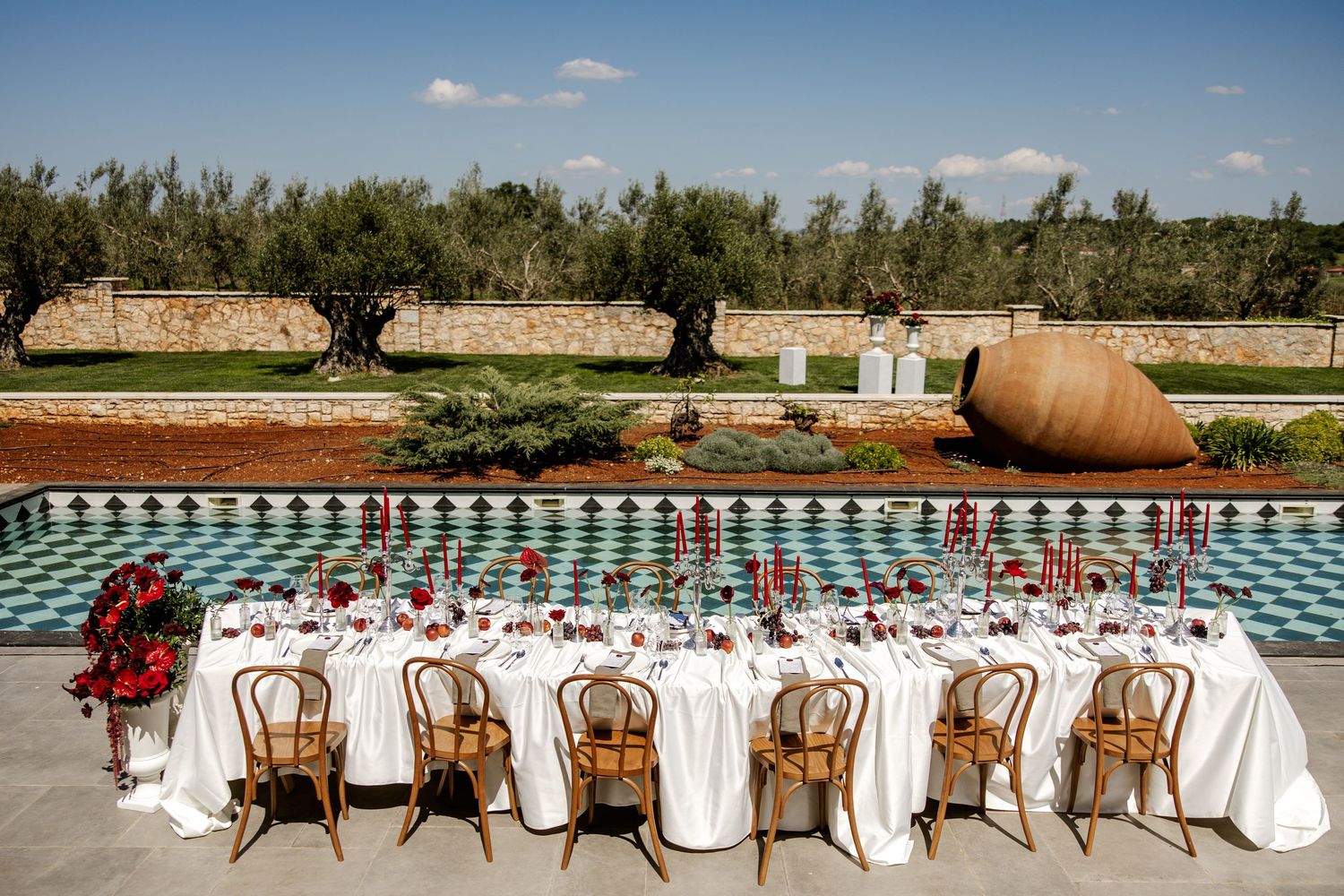 Elegant outdoor wedding reception table set poolside with white linens, red florals, and rattan chairs in a Mediterranean garden.