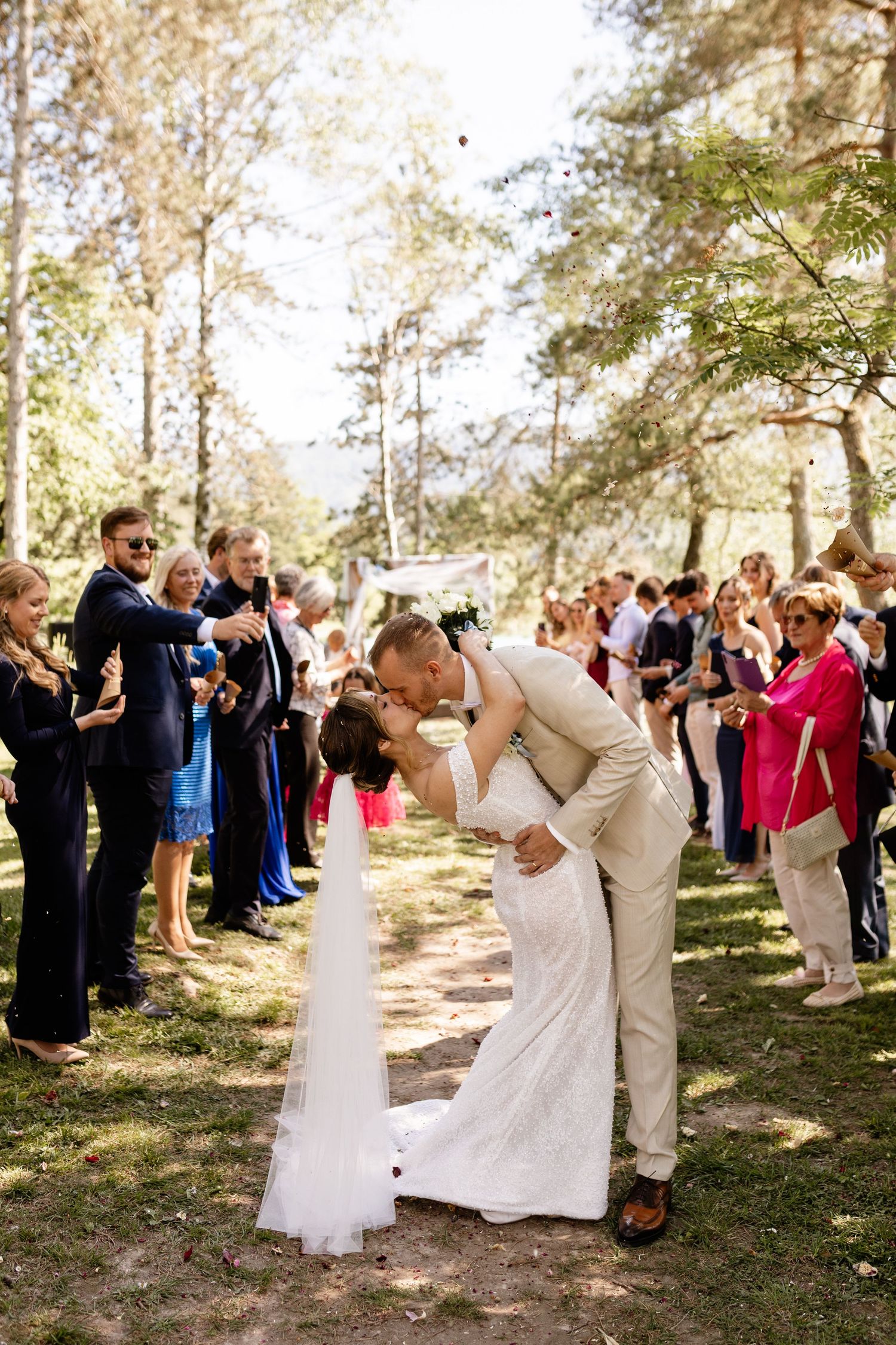 Bride and groom share a romantic dip kiss outdoors while wedding guests line the aisle throwing confetti.