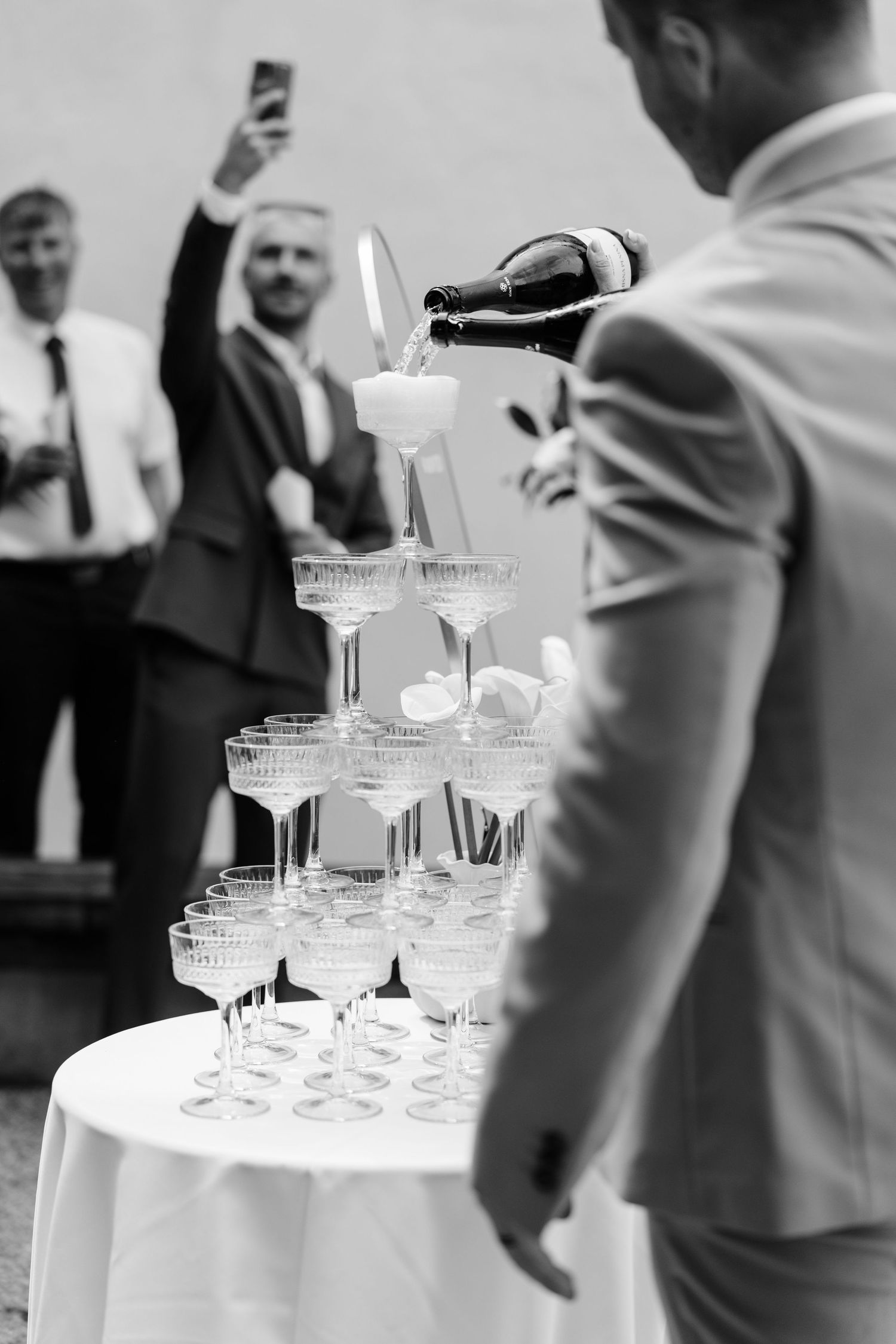 Groom pours champagne into elegant tower of glasses at wedding reception in black and white.