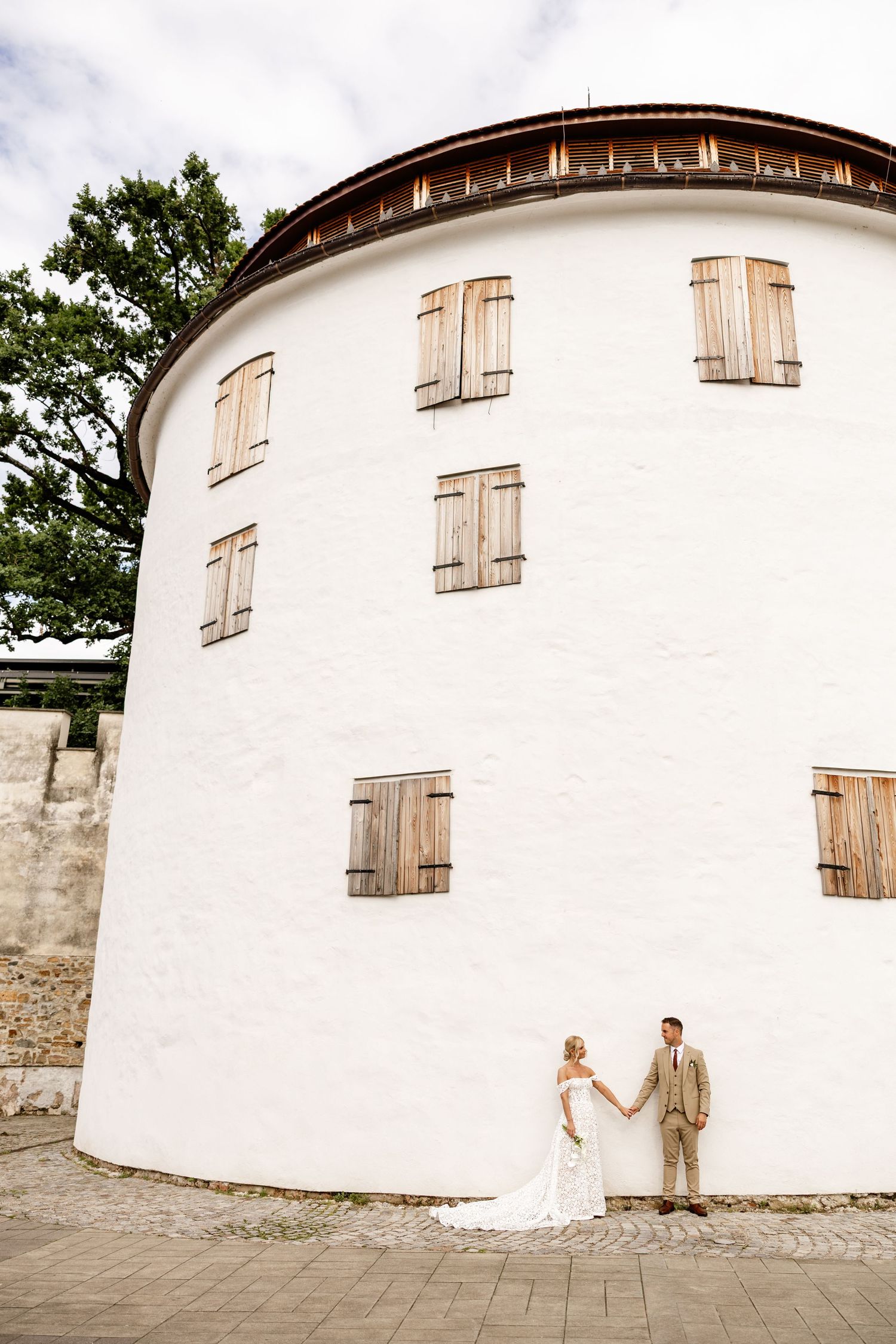 Wedding couple holds hands in front of a curved white building with wooden shuttered windows on cobblestone.