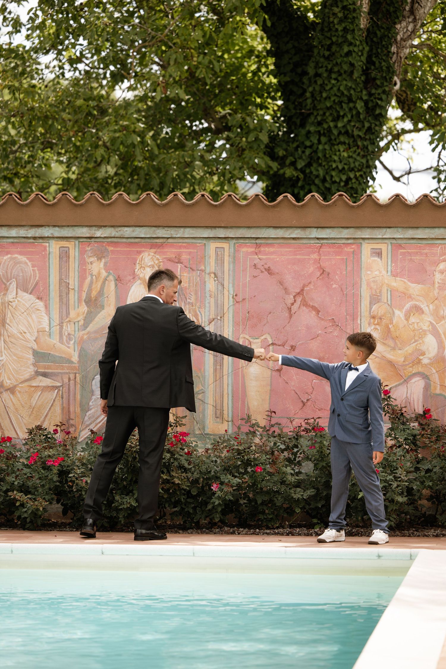 Groom in suit shaking hands with young boy in bow tie beside outdoor pool.