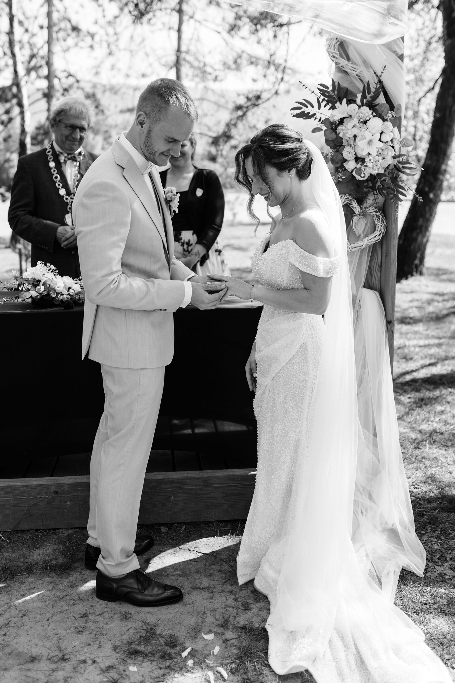 Black and white photo of groom placing wedding ring on brides finger during outdoor ceremony.