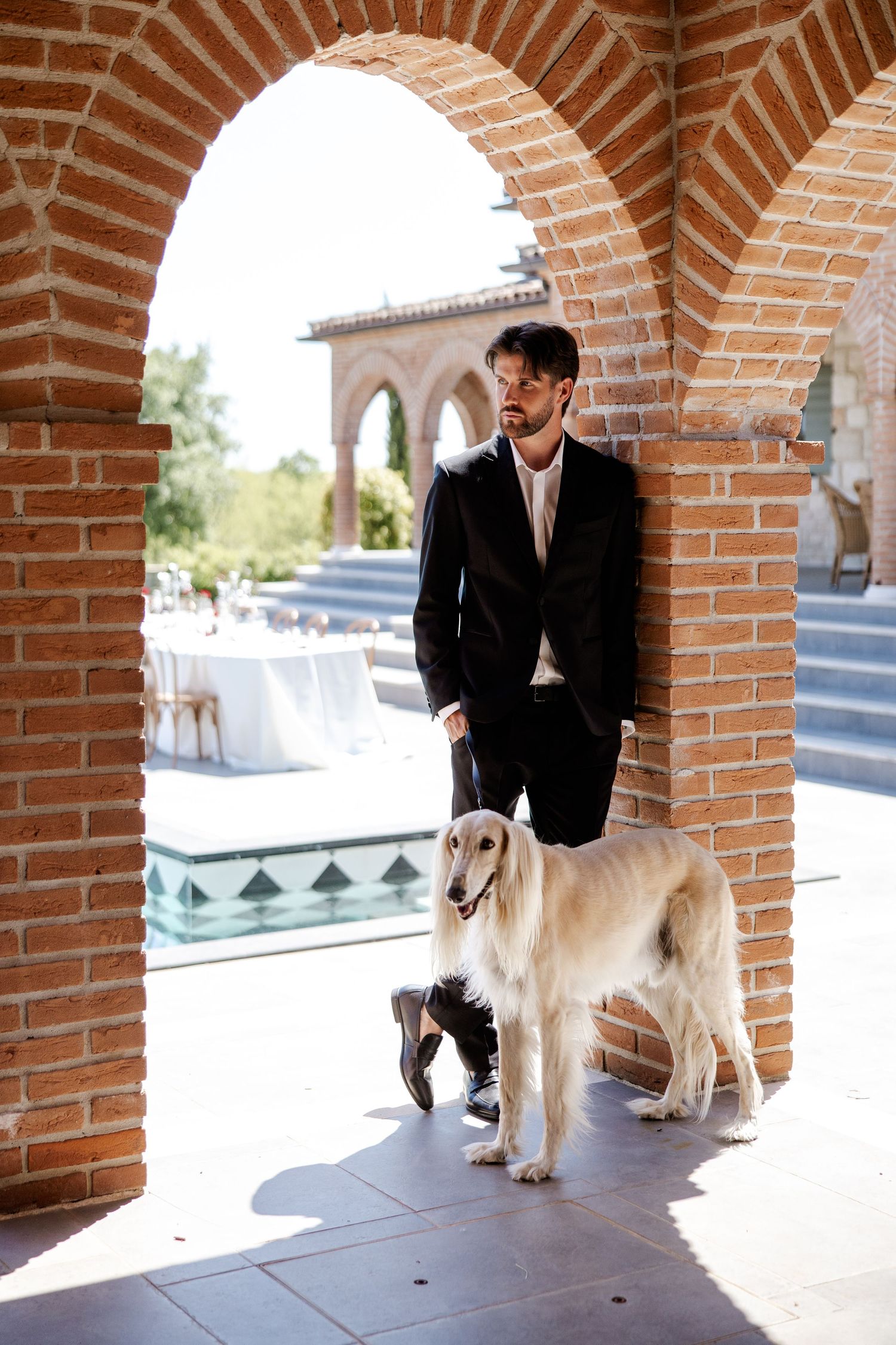 Groom in black suit stands with elegant white Saluki dog under brick arches at outdoor wedding venue.