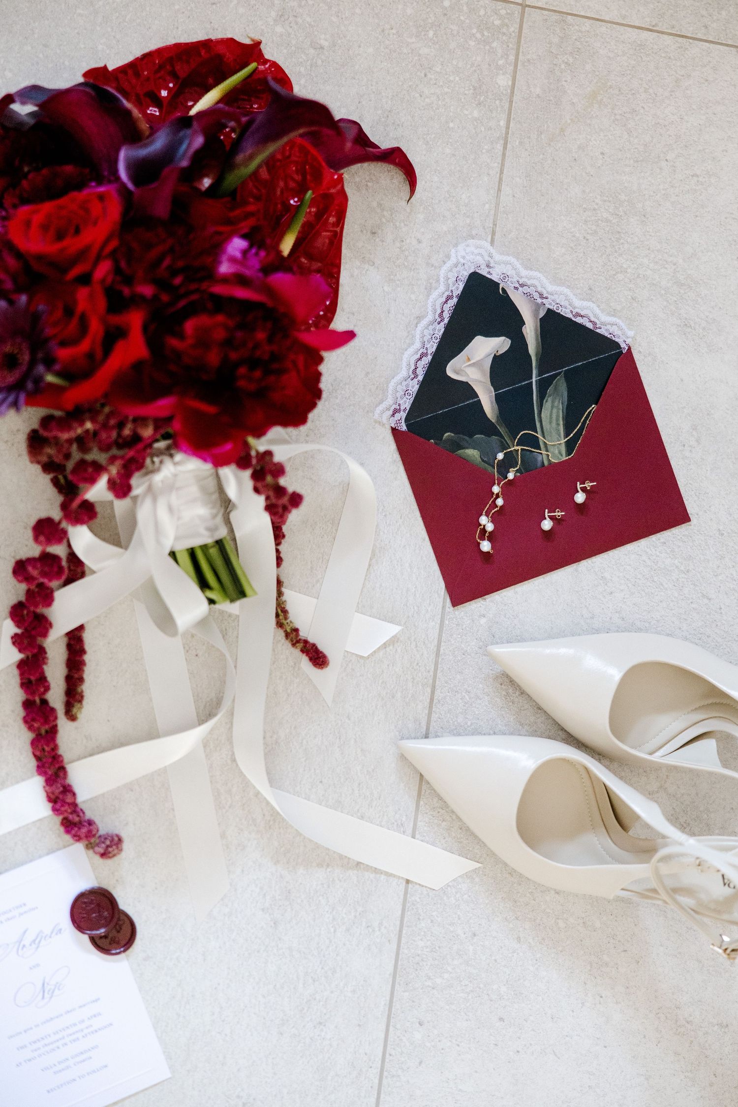 Bridal flat lay with deep red bouquet, white heels, burgundy envelope, earrings, and wedding invitation on tile floor.