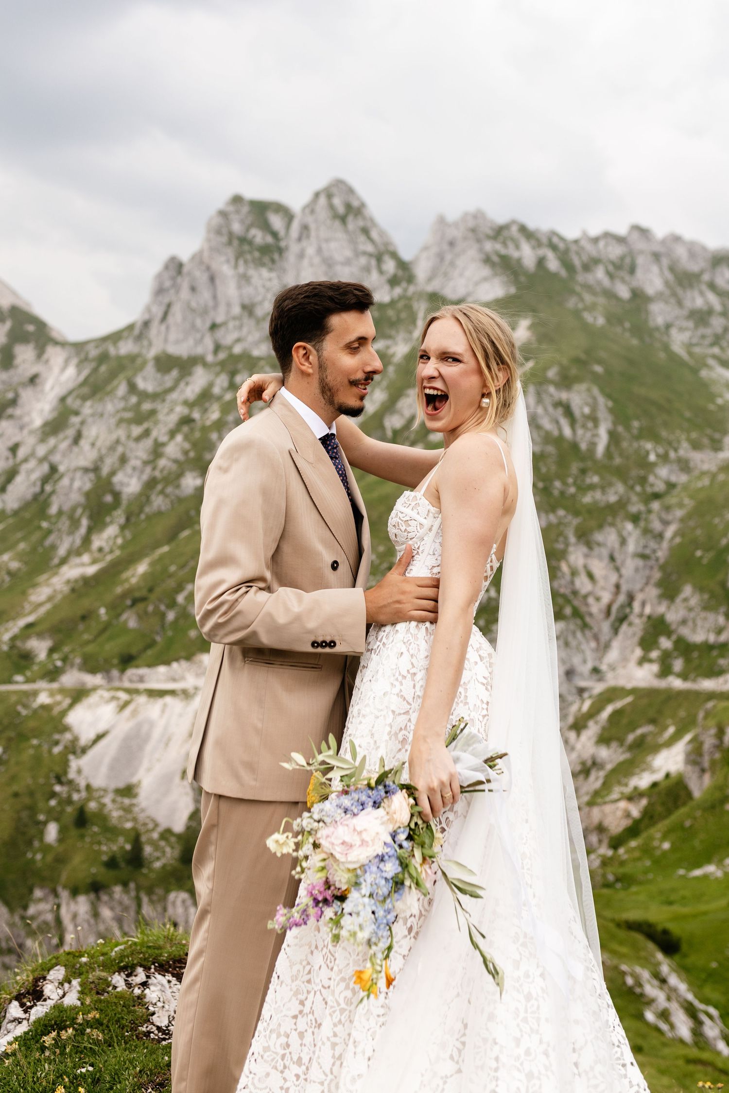 Joyful bride in lace gown with floral bouquet and groom in tan suit embracing before Alpine mountain backdrop.