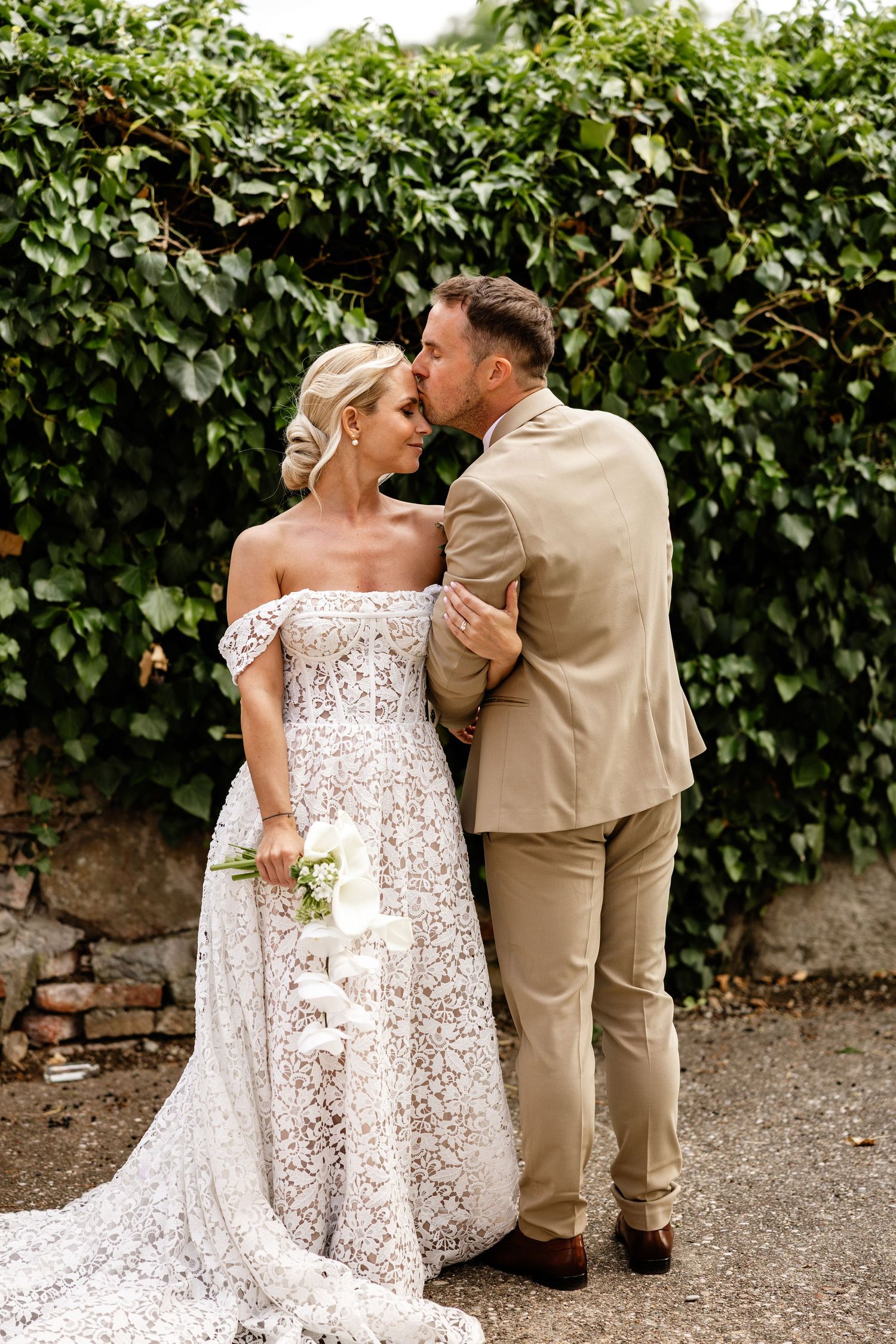 Bride and groom share a romantic kiss outdoors, with lush green ivy wall backdrop, bride holding white bouquet.