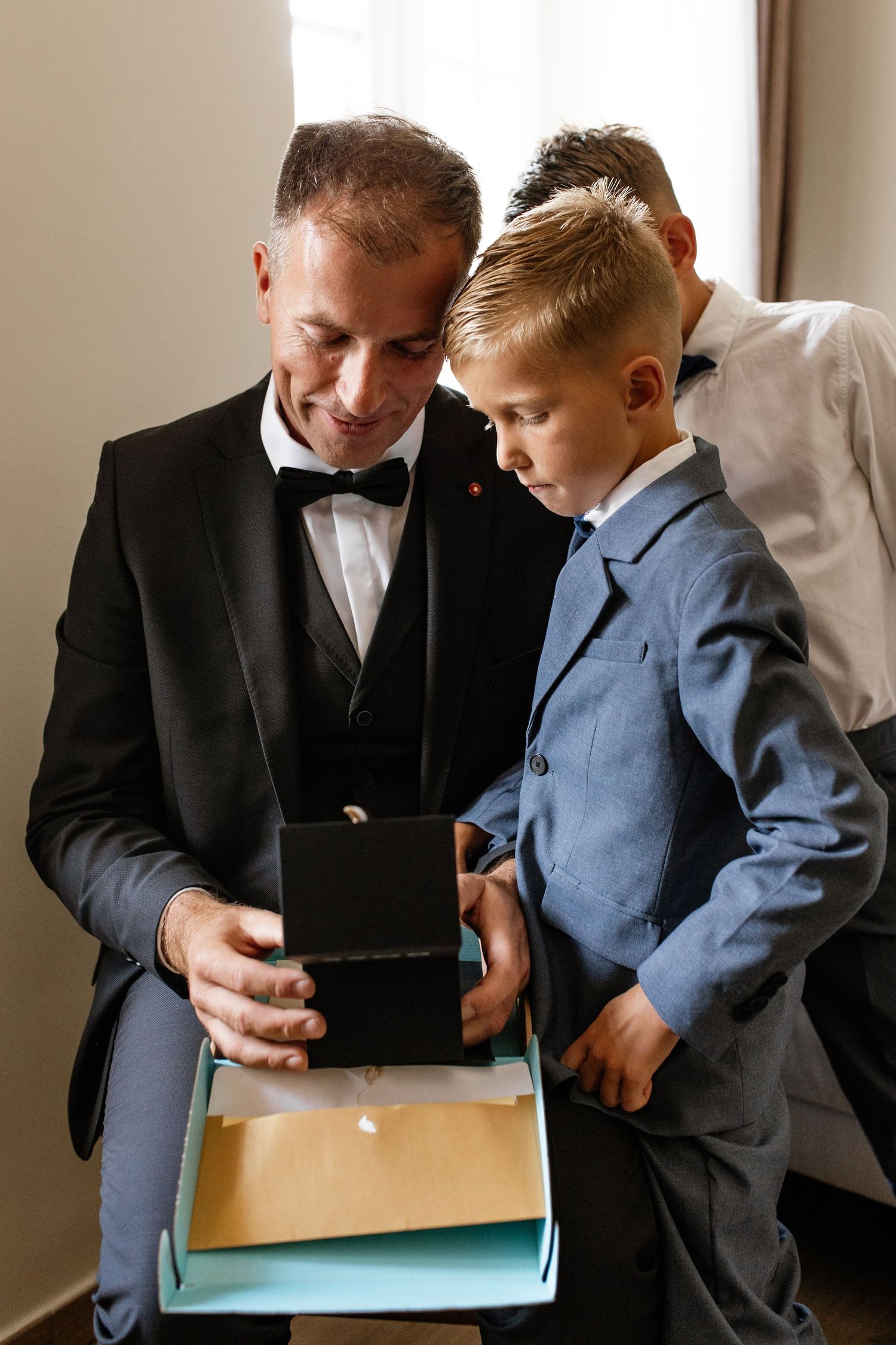 Man in tuxedo and boy in blue suit examining gift box together at formal event.