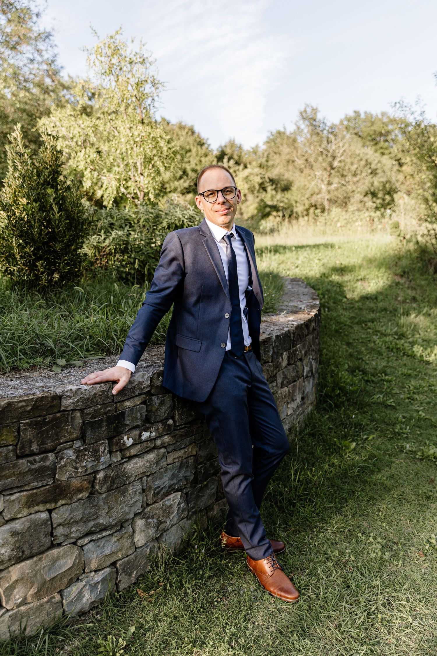 Stylish groom in a navy blue suit and brown shoes leaning against a stone wall in a lush green outdoor setting.