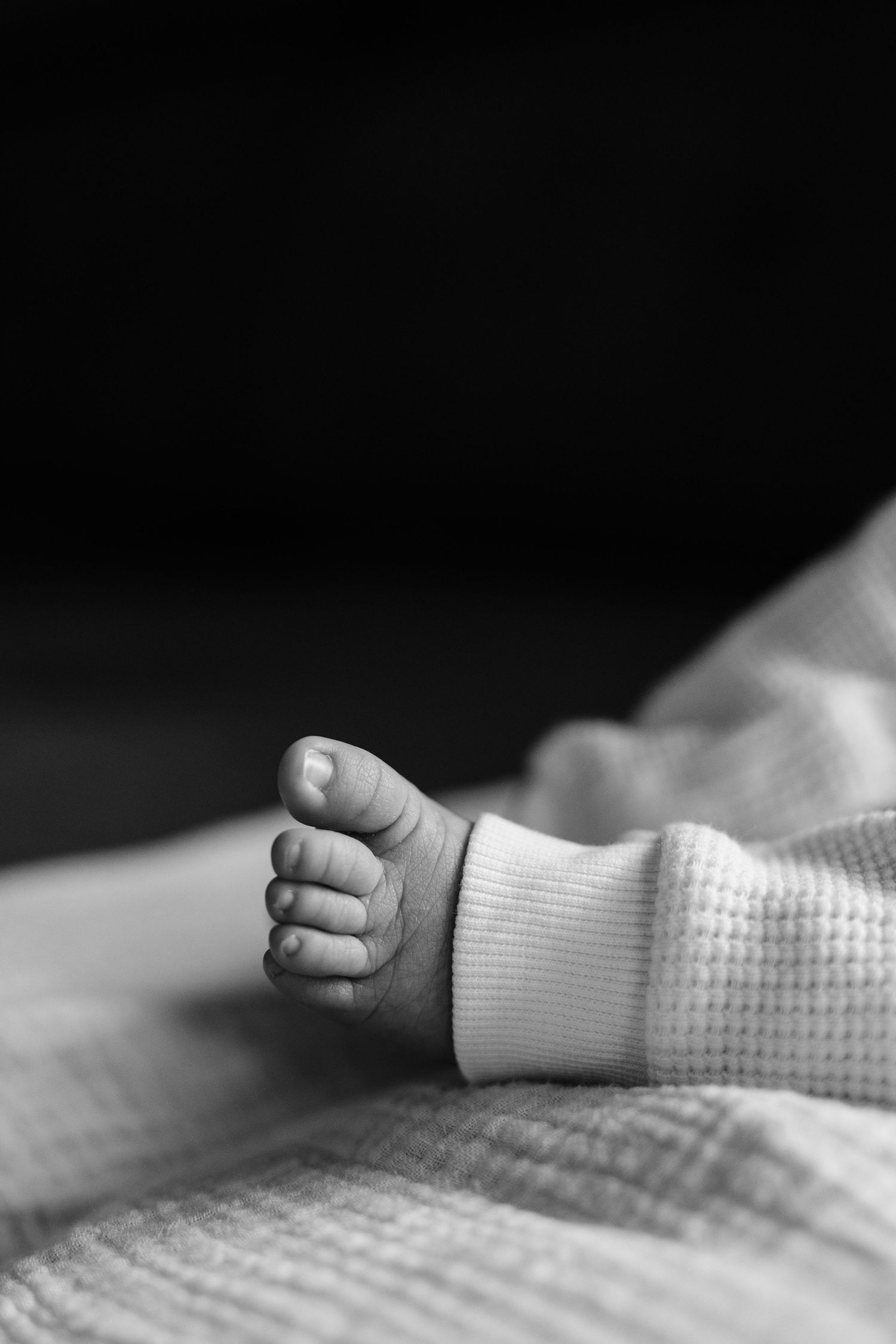 Black and white close-up photo of a newborn baby's tiny foot in a knit onesie resting on a soft blanket.