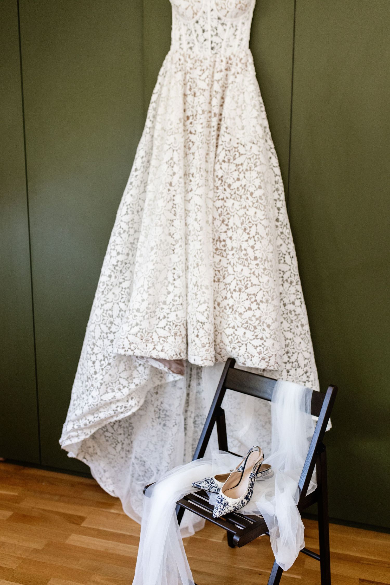 Elegant white lace wedding dress hanging above a chair with bridal heels and veil in front of olive green wall.