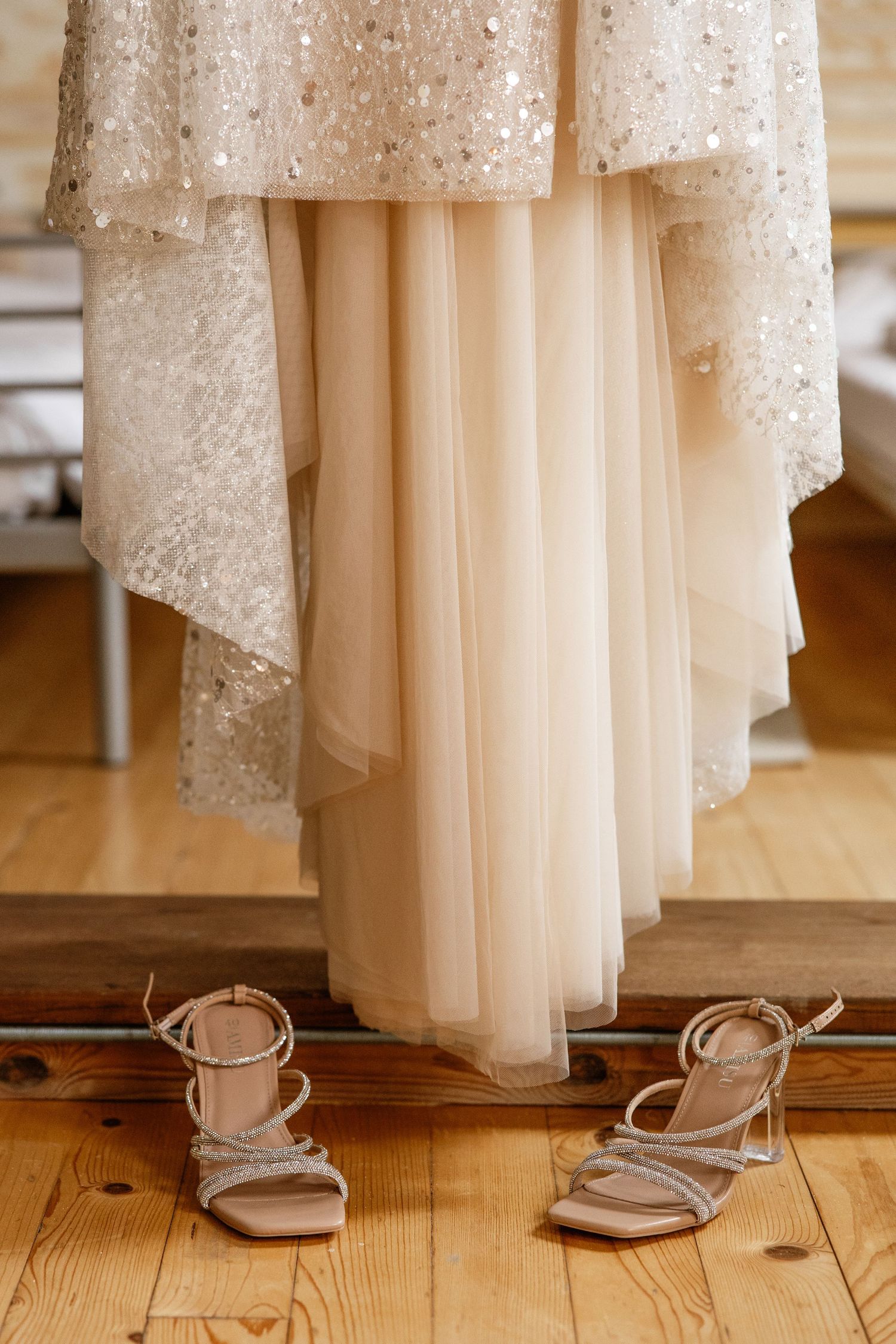 Beaded ivory wedding dress hanging above strappy crystal bridal heels on wood floor.