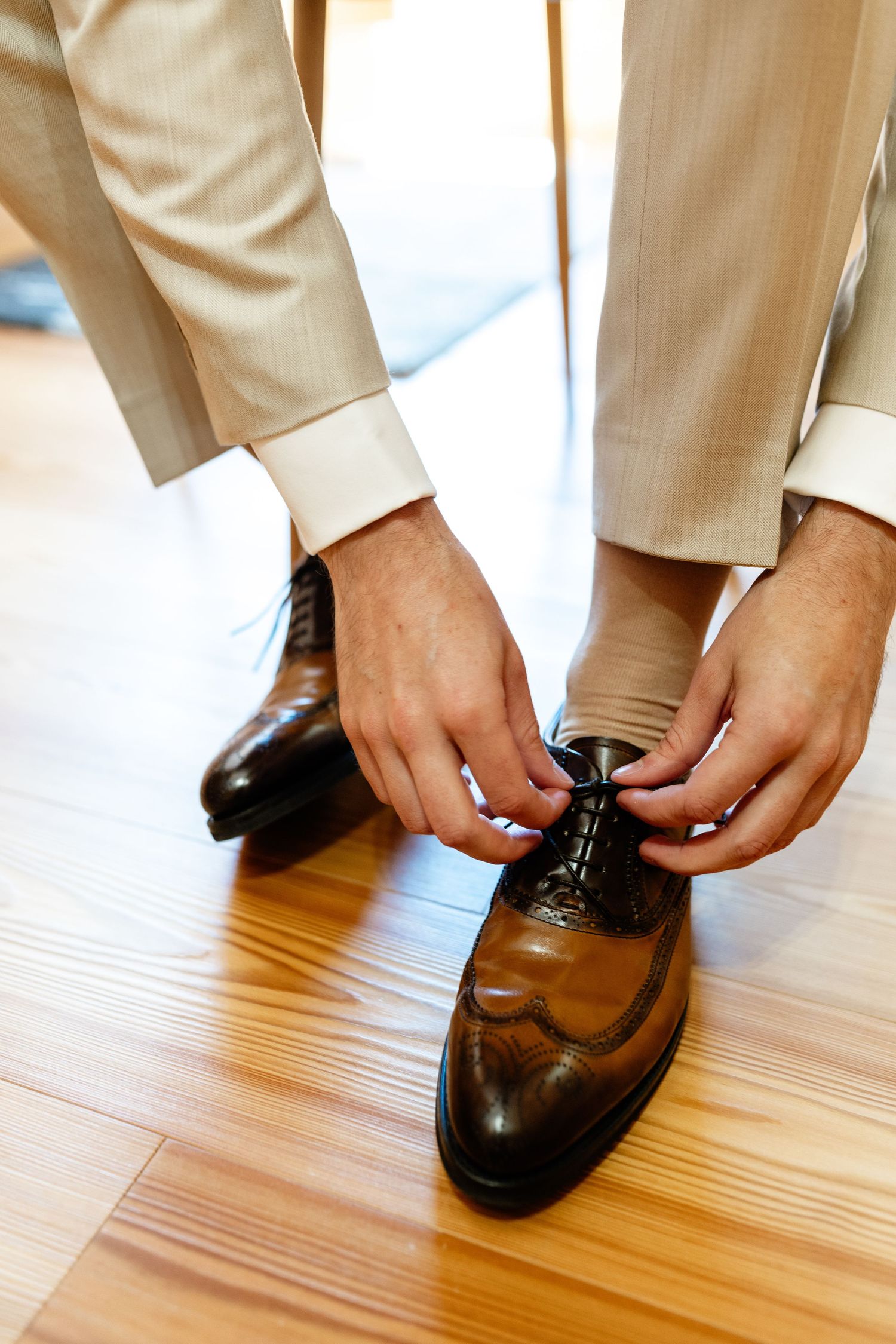 Man in beige suit tying brown leather oxford brogue dress shoes on hardwood floor.