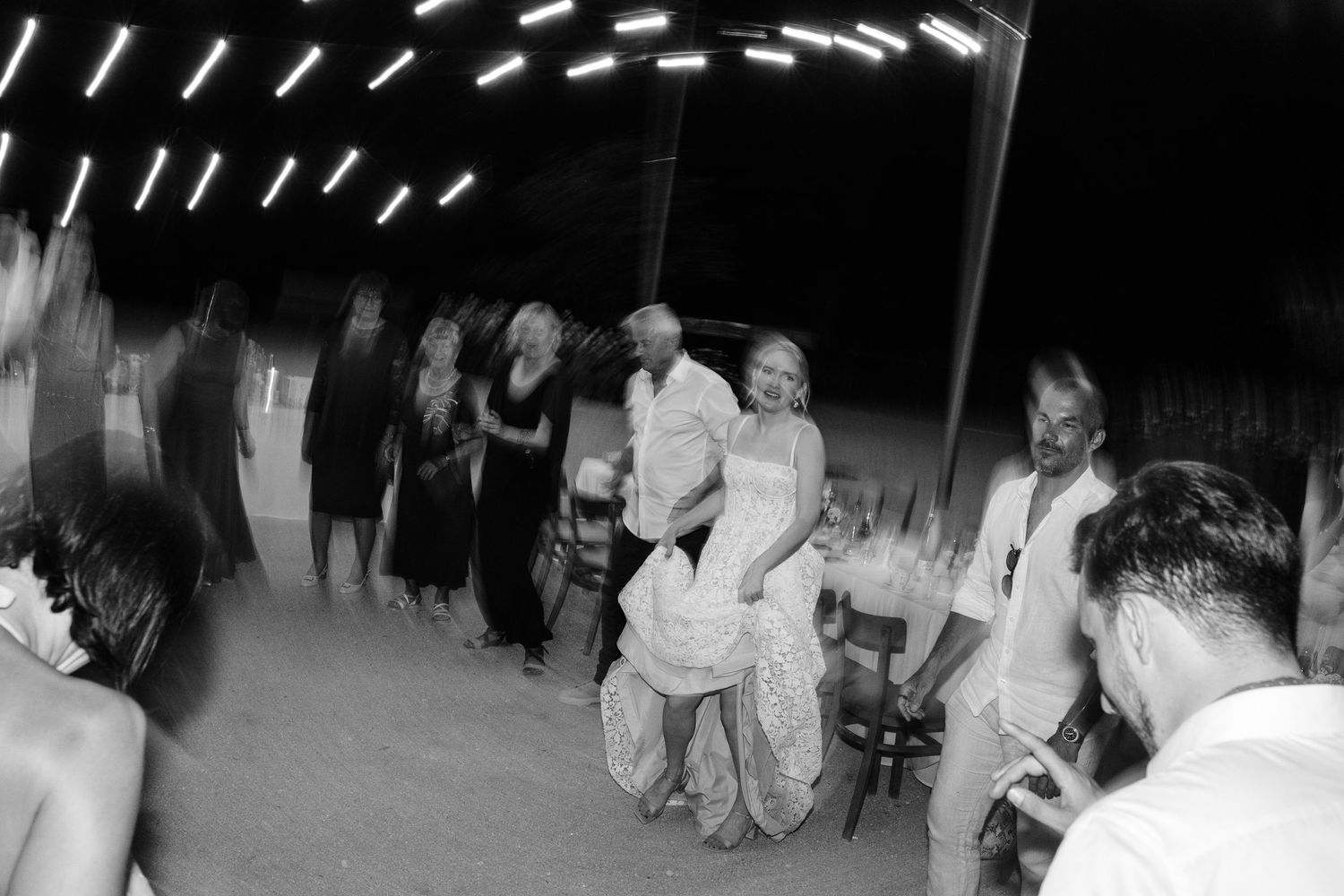 Black and white photo of bride and groom dancing with guests at an outdoor wedding reception under string lights.