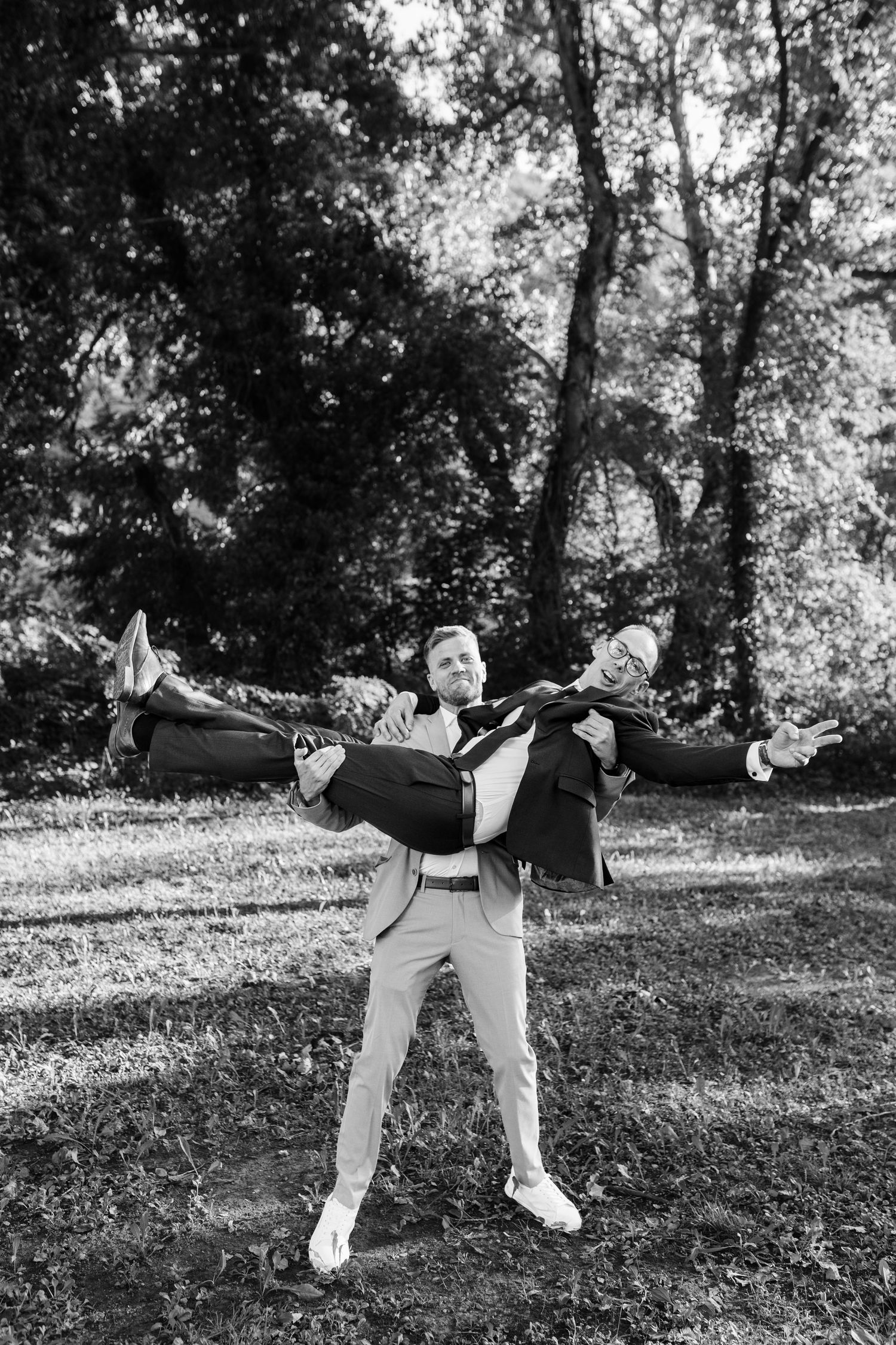 Two groomsmen celebrate outdoors as one carries the other in a playful black and white photo.