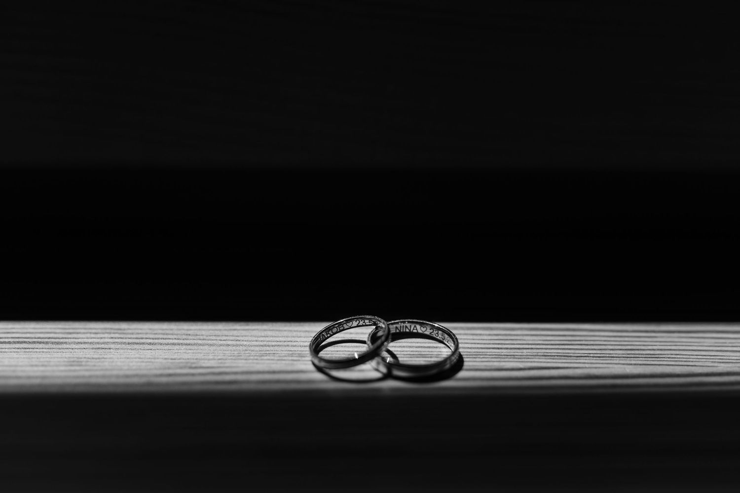 Two elegant wedding rings resting together on a wooden surface in a dramatic black and white photograph.