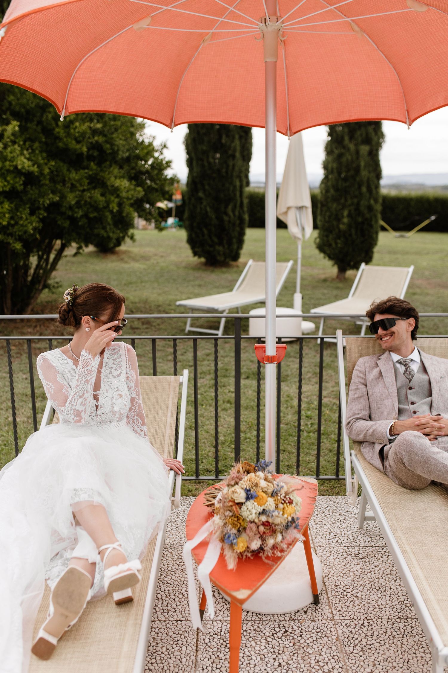 Bride and groom relax on sun loungers under an orange umbrella with a dried floral bouquet between them.