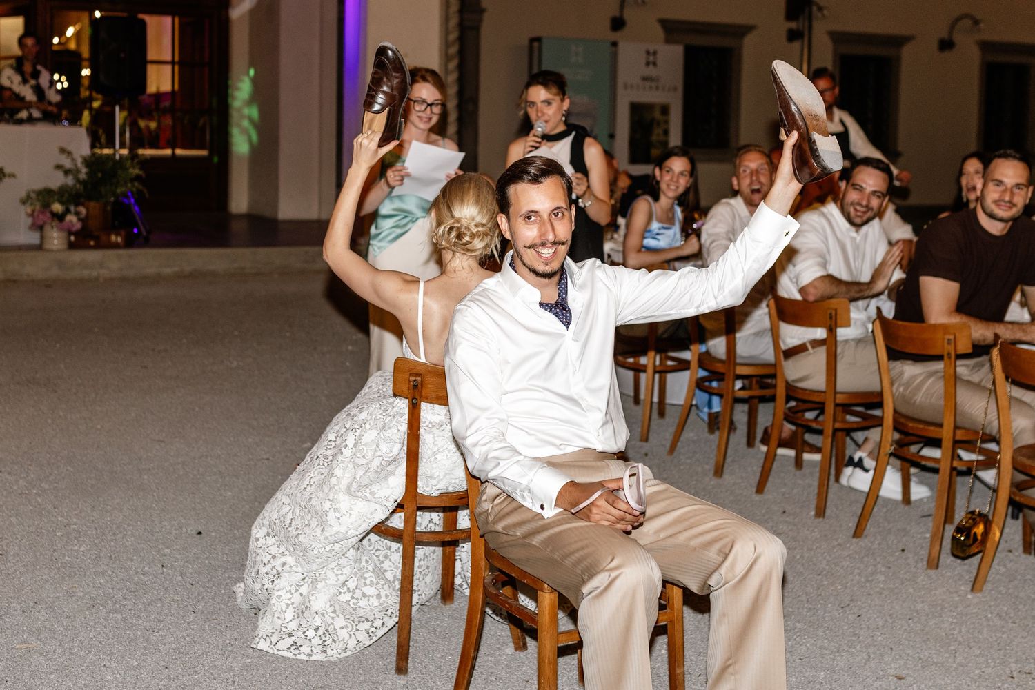 Bride and groom sit on chairs raising hands at outdoor wedding reception with guests in background at night.