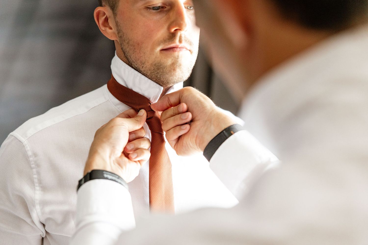 Man in white dress shirt getting his orange tie adjusted by another person wearing a black watch.