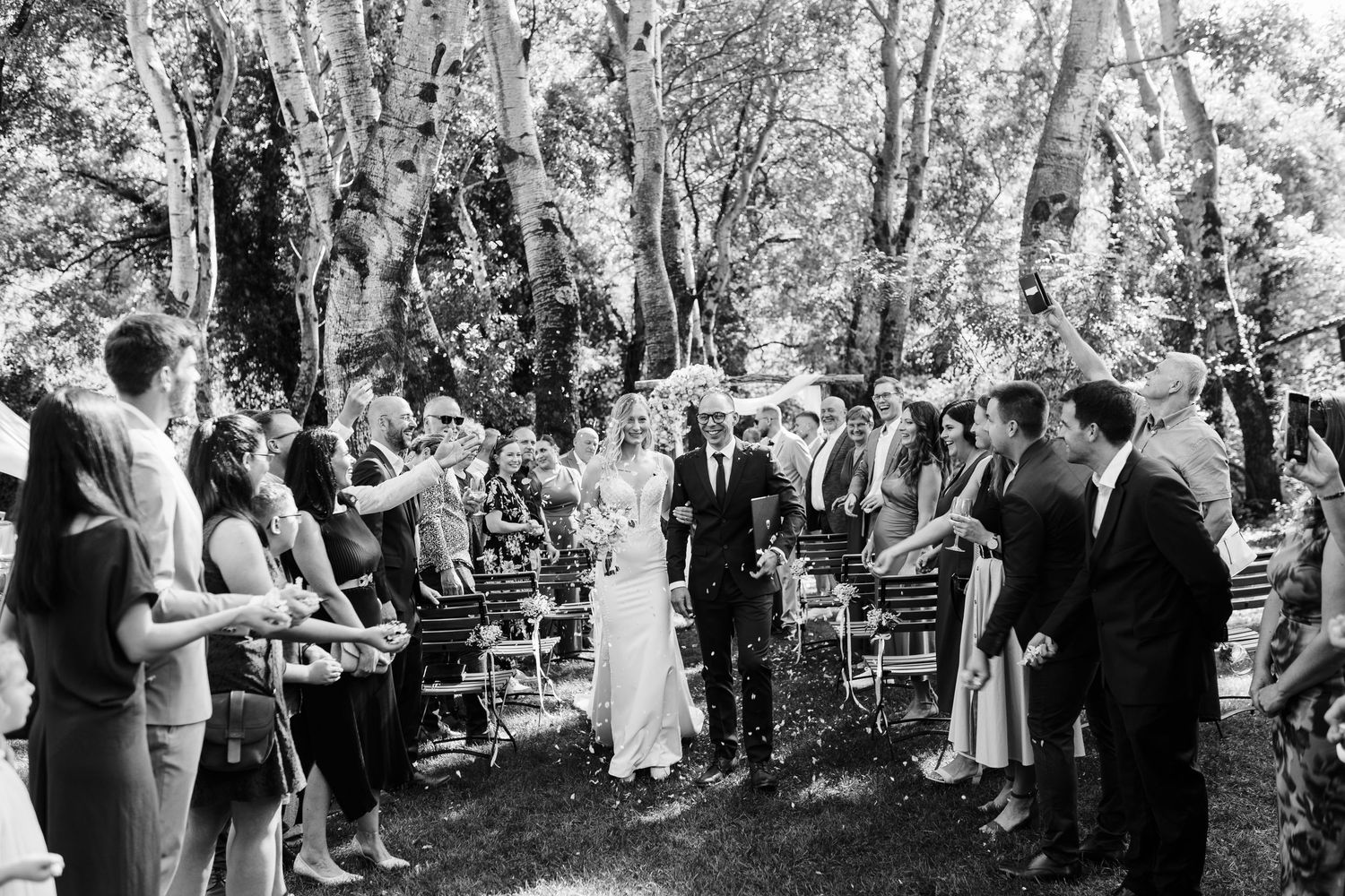 Black and white outdoor wedding ceremony with couple walking down aisle surrounded by guests and tall trees.