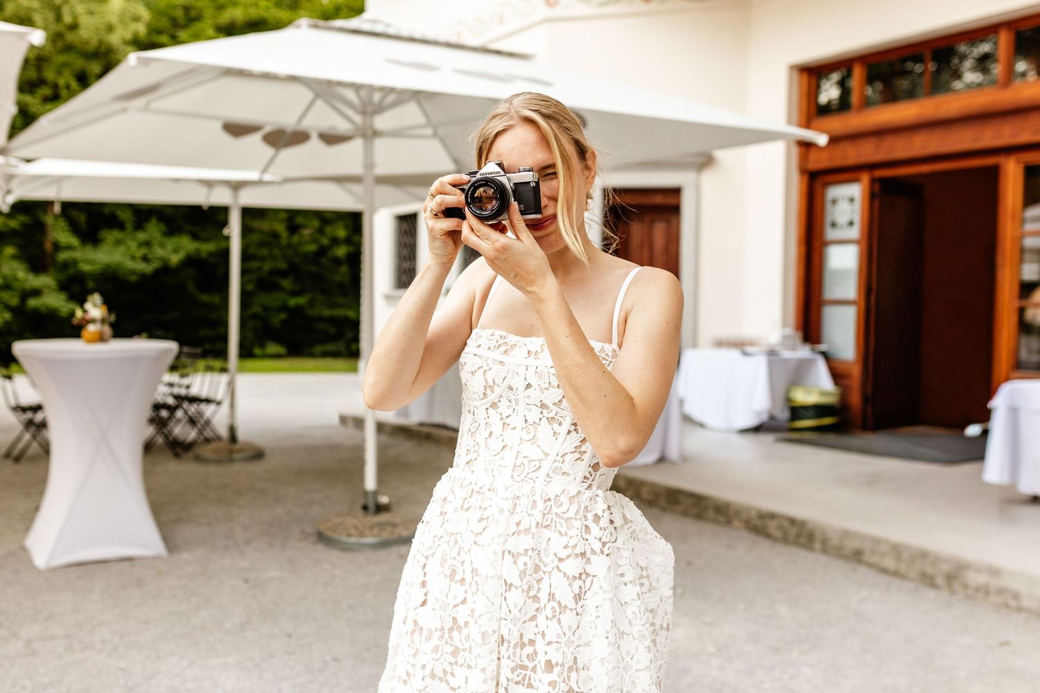 Blonde woman in white lace dress photographing with vintage film camera at outdoor wedding venue.