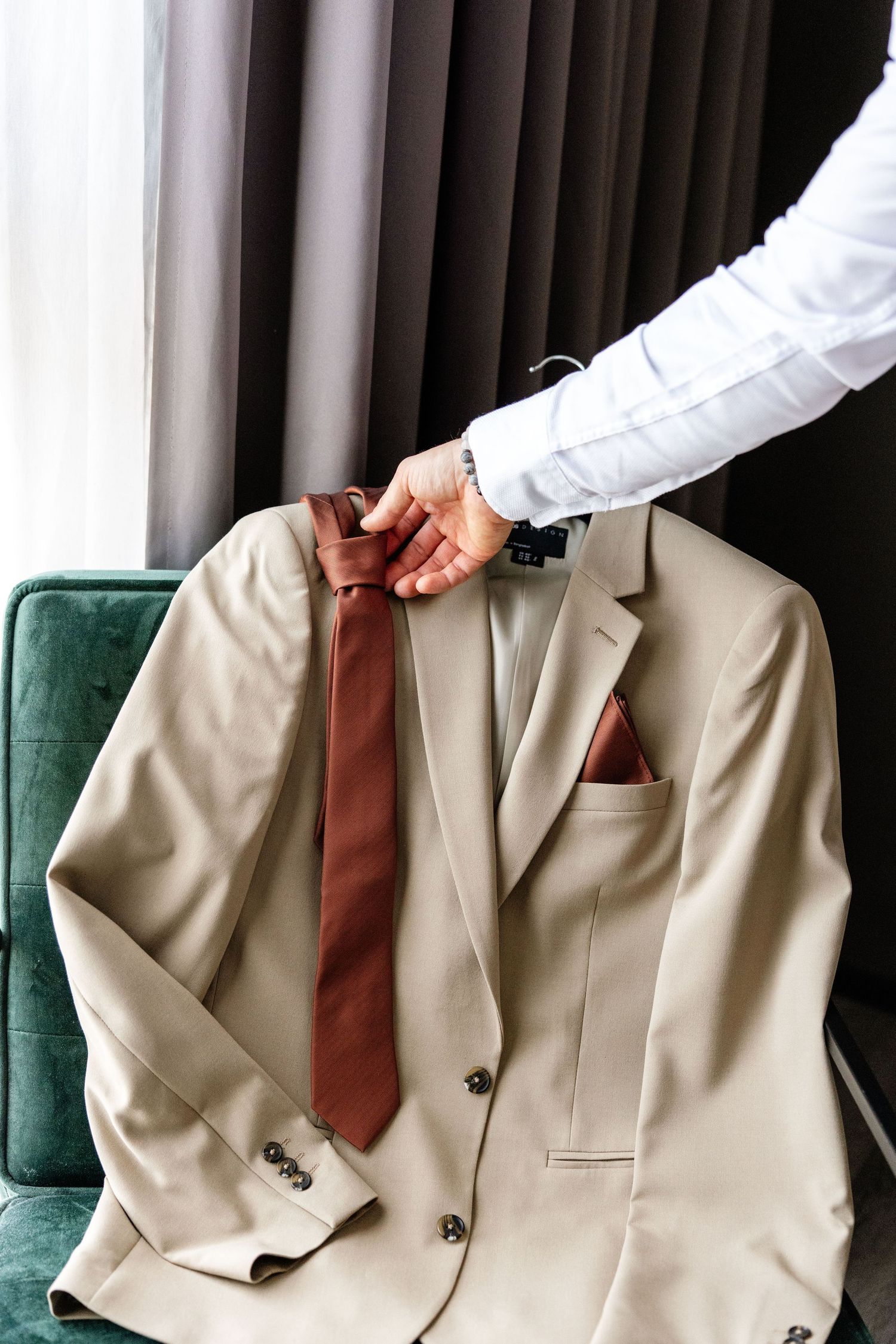 Groom's hand adjusting brown silk tie on beige wedding suit with pocket square on green velvet chair.