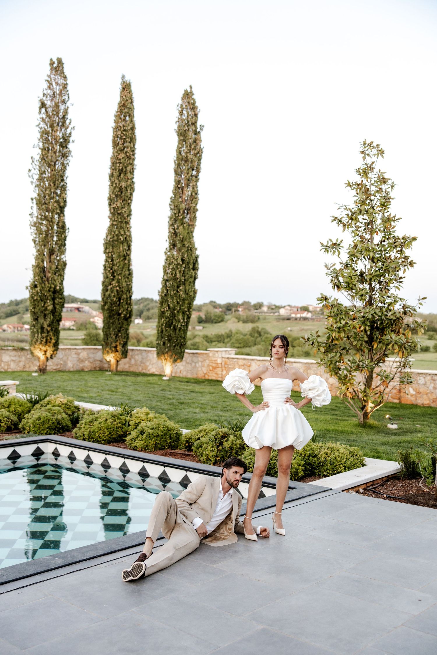 Bride in white puff-sleeve mini dress standing poolside while groom reclines on the ground near cypress trees.