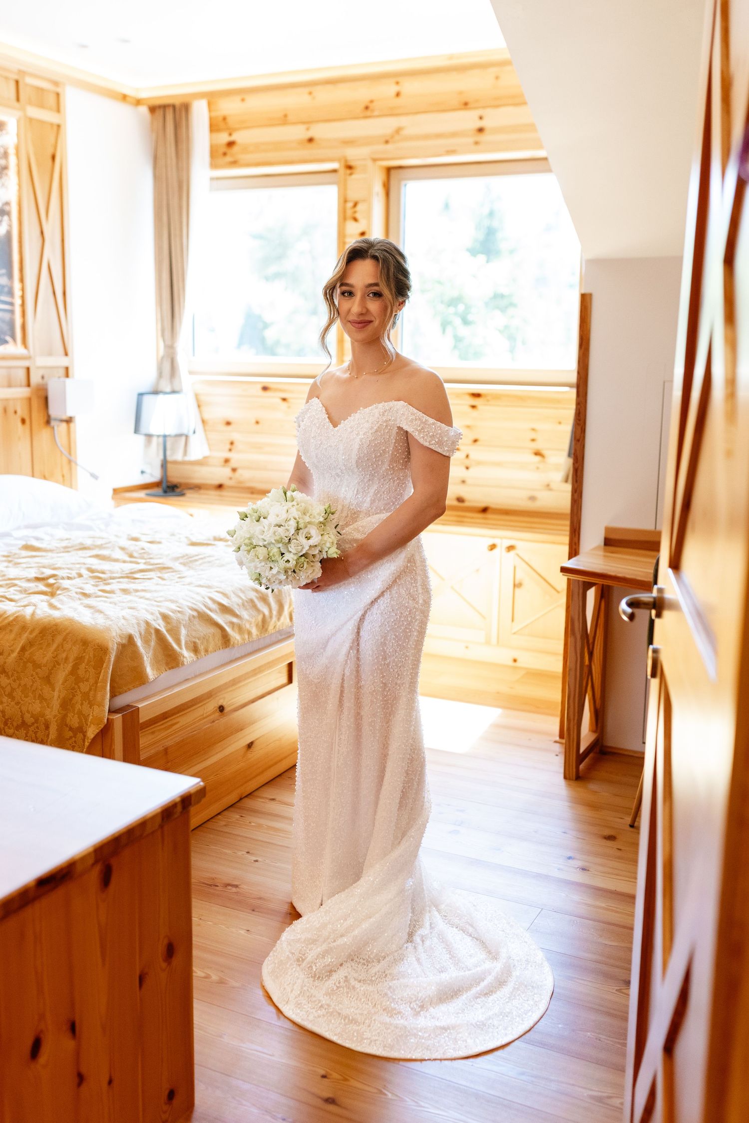 Beautiful bride in a fitted off-shoulder lace wedding gown holding a bouquet in a rustic wooden room.