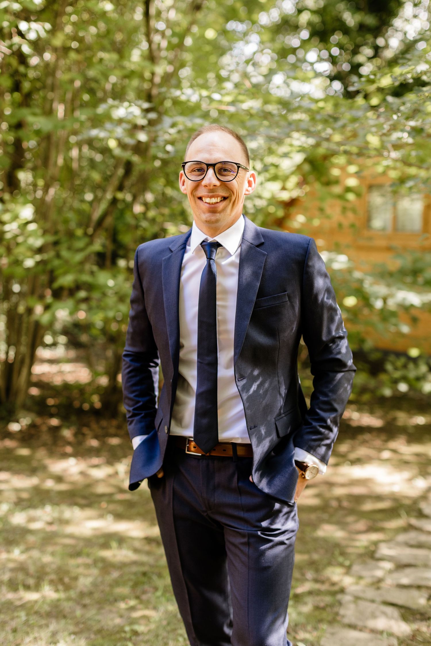 Smiling groom in navy blue suit and tie with glasses standing outdoors among green trees.