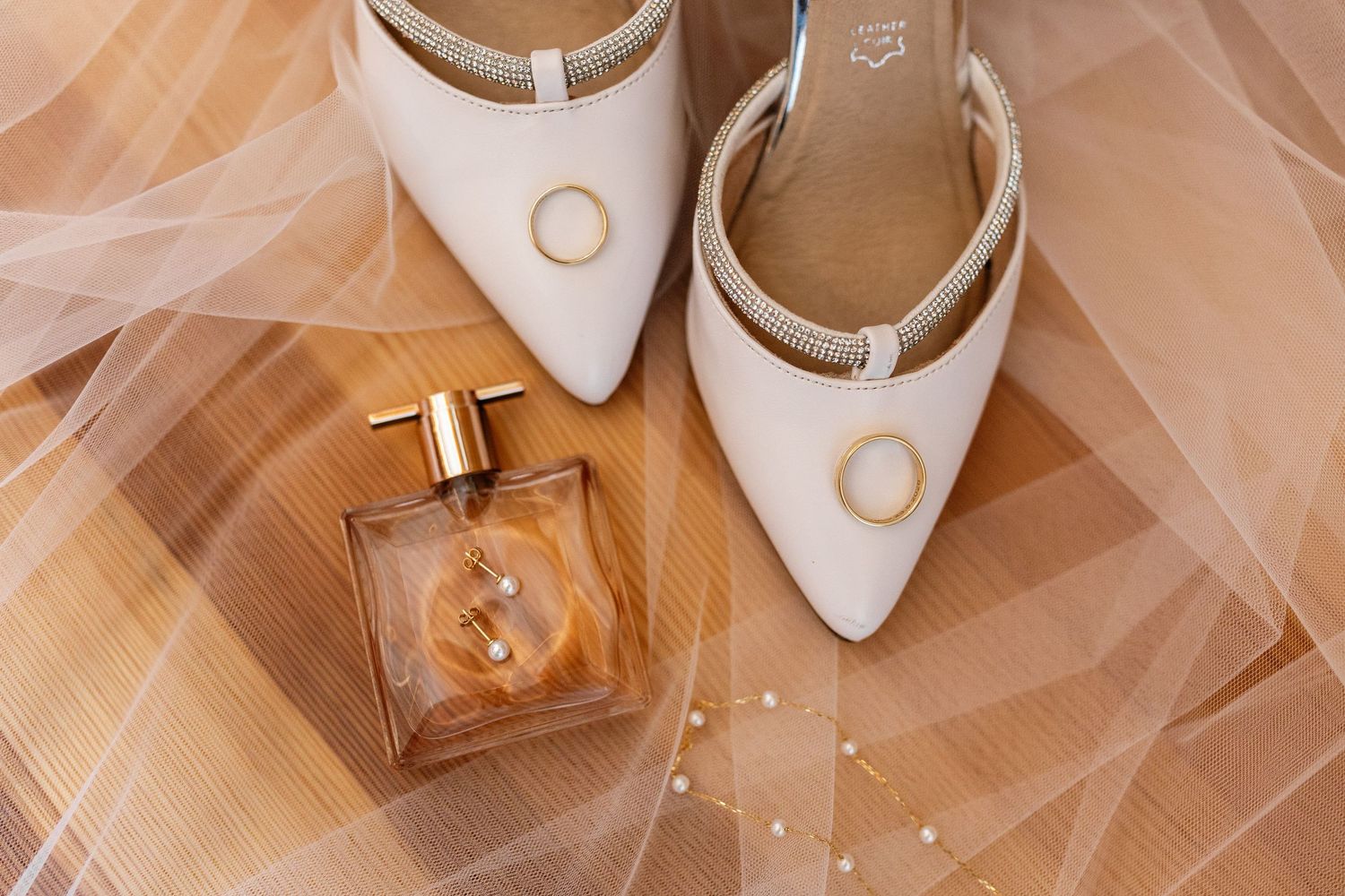 Elegant bridal accessories featuring white pointed-toe heels, gold wedding rings, and amber perfume bottle on sheer veil.