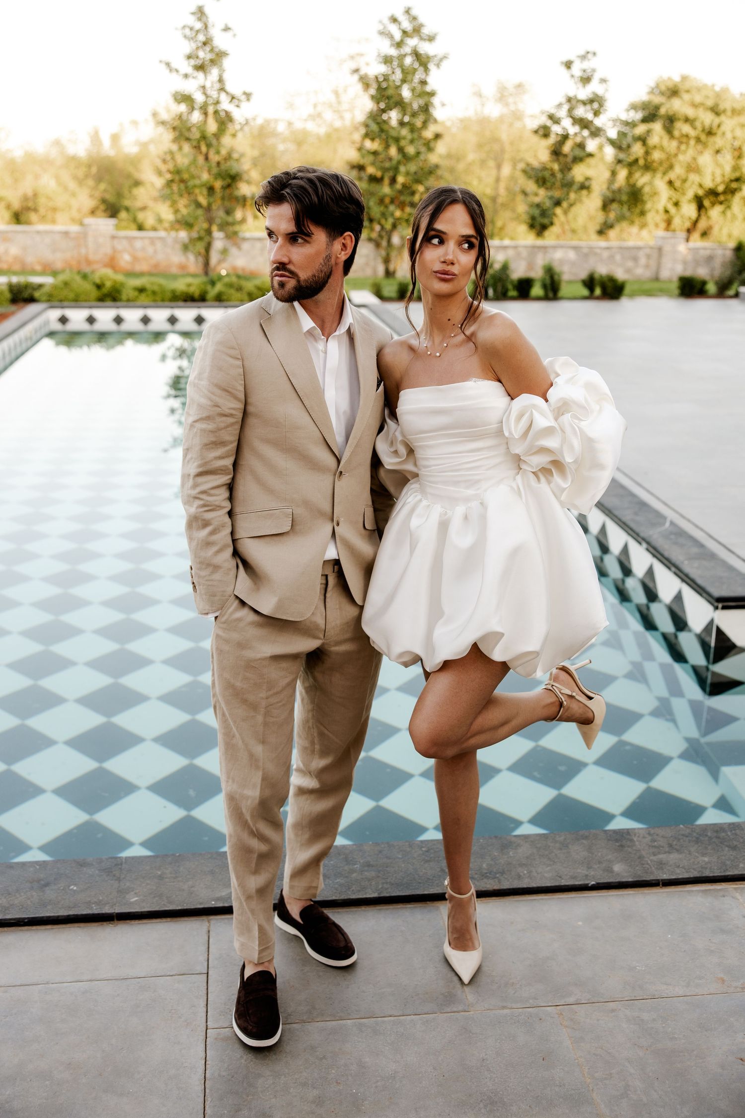 Elegant couple posing by a checkered pool, groom in beige suit and bride in a white strapless puff-sleeve mini dress.