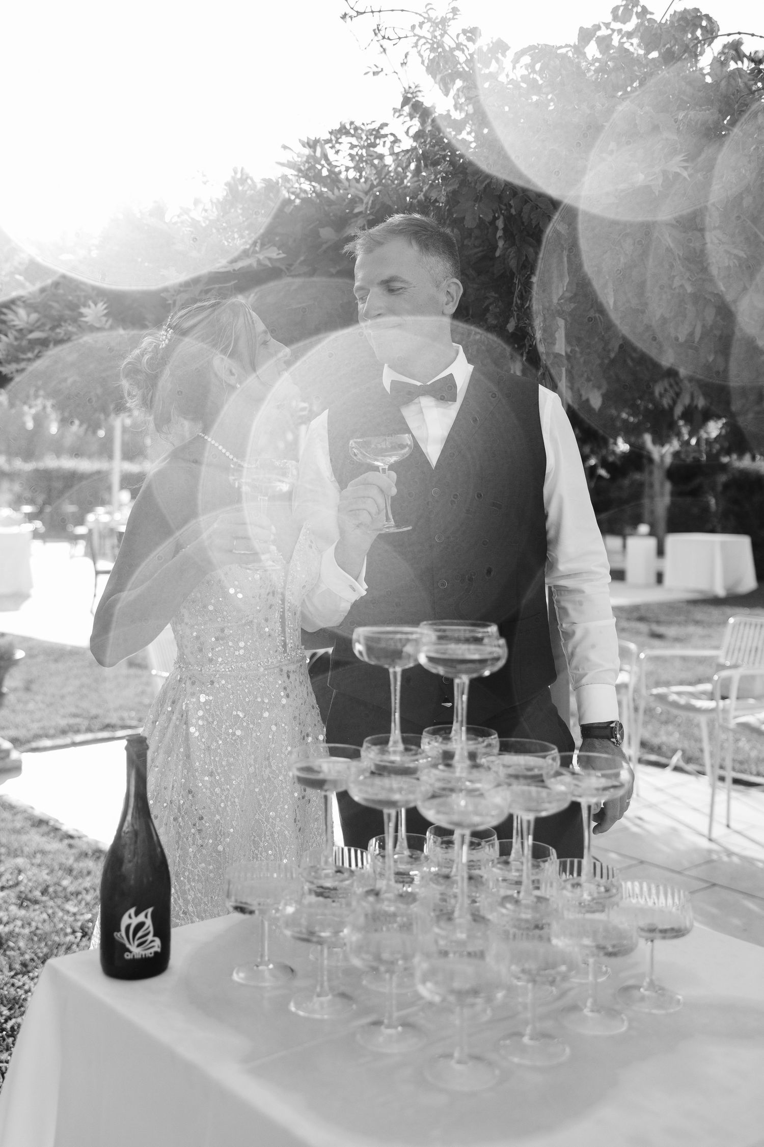 Black and white wedding photo of couple kissing beside champagne tower and bottle at outdoor reception.