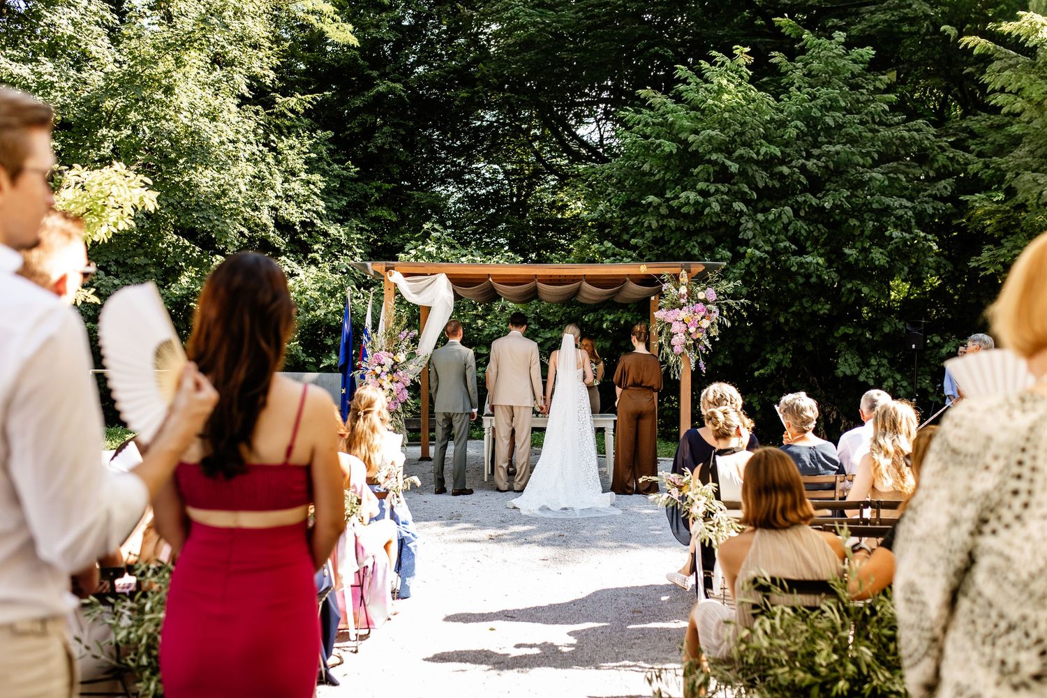 Outdoor wedding ceremony with guests watching a couple exchange vows beneath a floral arch surrounded by trees.