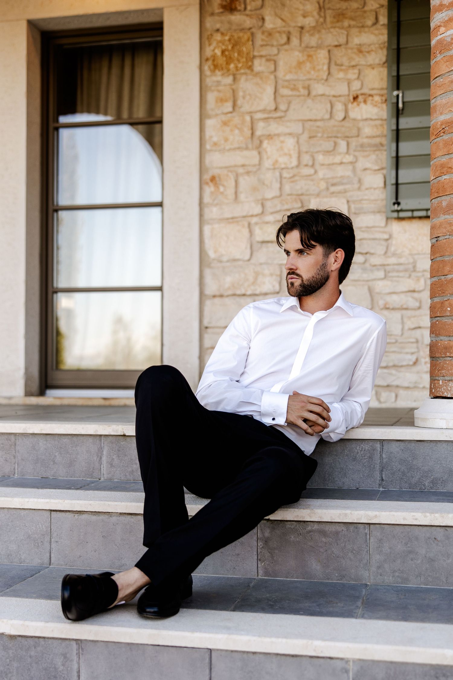 Man in white dress shirt and black pants sitting on stone steps outside a building, looking away thoughtfully.