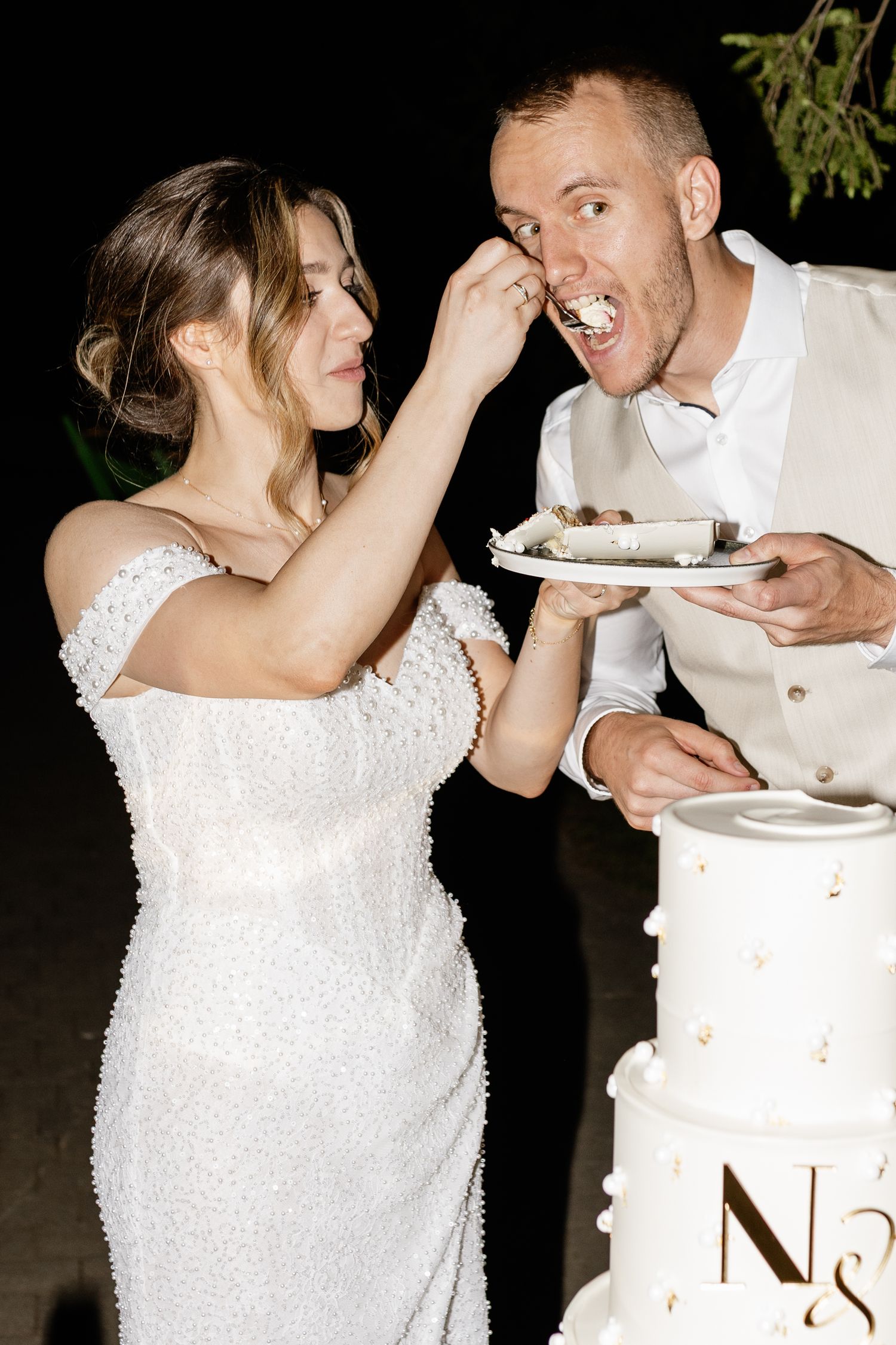 Bride playfully feeds wedding cake to her groom beside a white tiered cake at their night reception.