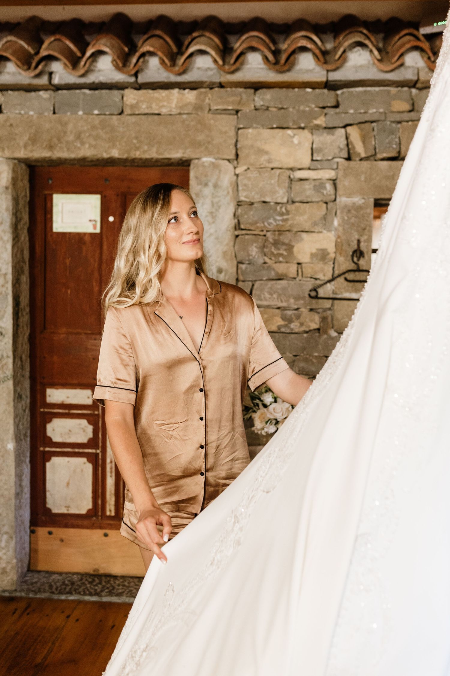 Bride in champagne satin pajamas admiring her wedding dress against a rustic stone wall backdrop.
