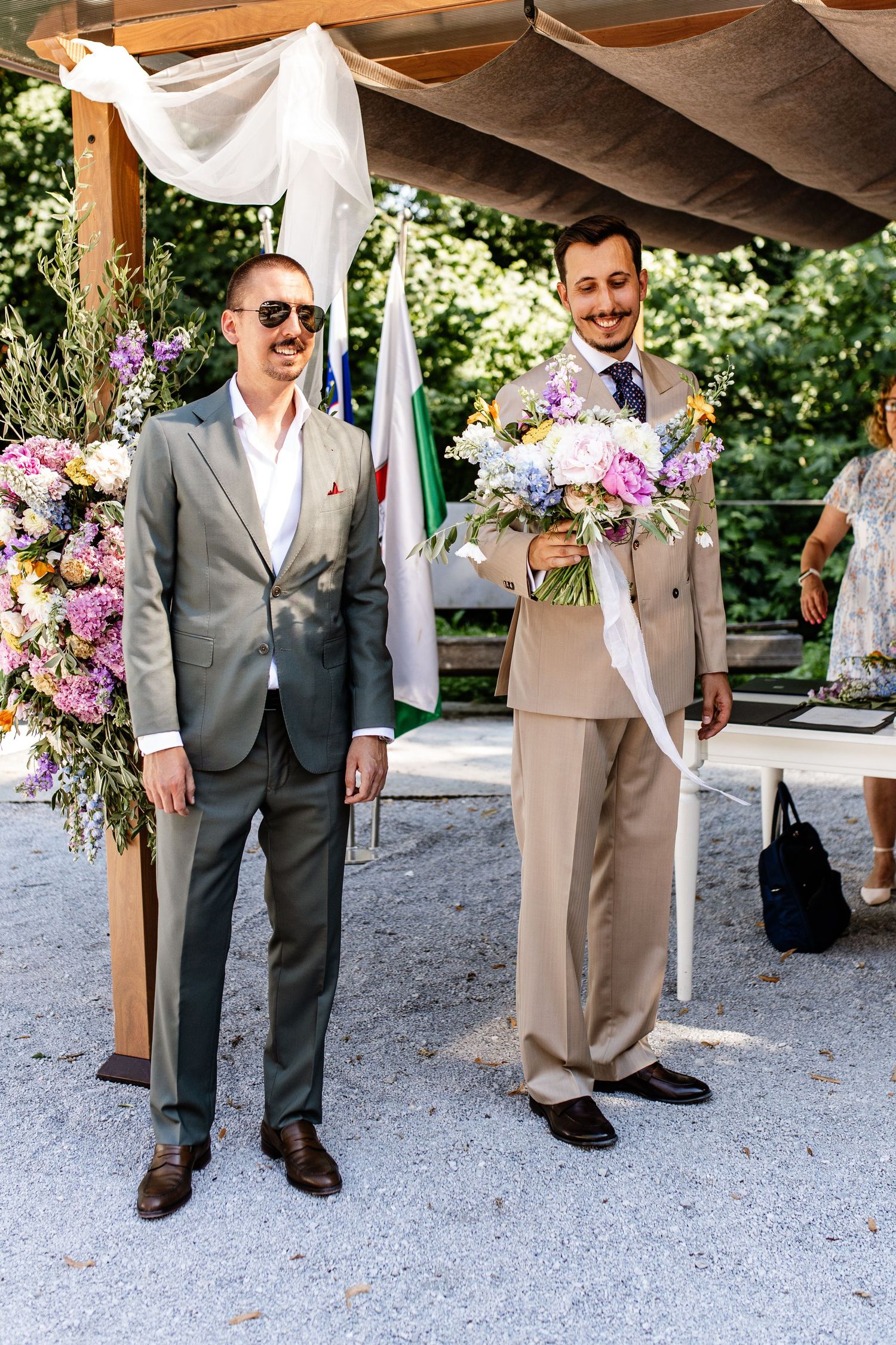 Two grooms smile at outdoor wedding ceremony, one holding colorful bouquet, both wearing suits under floral decorated arbor.