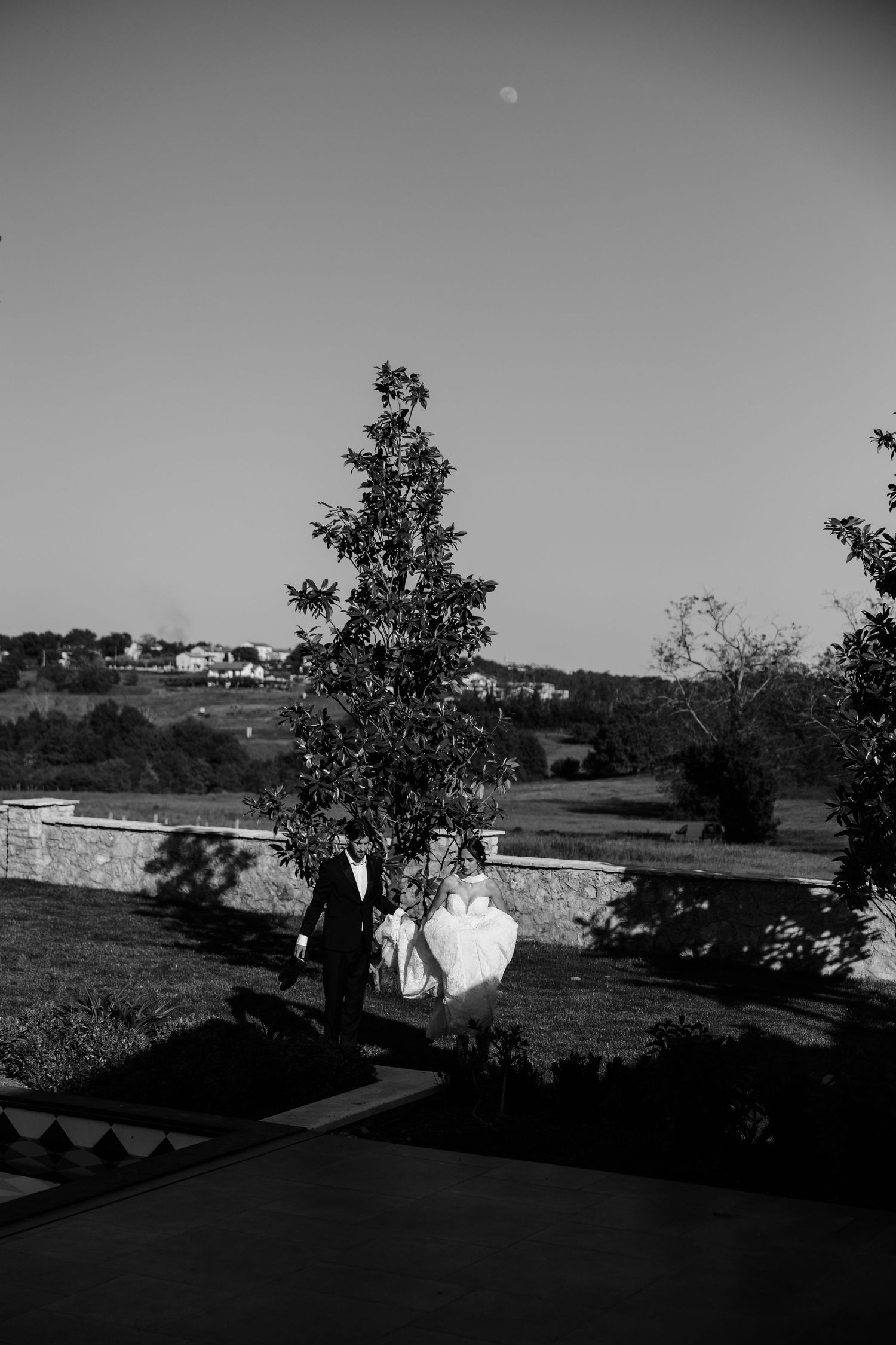 Black and white wedding photo of bride and groom walking outdoors under moonlit sky near a tree.