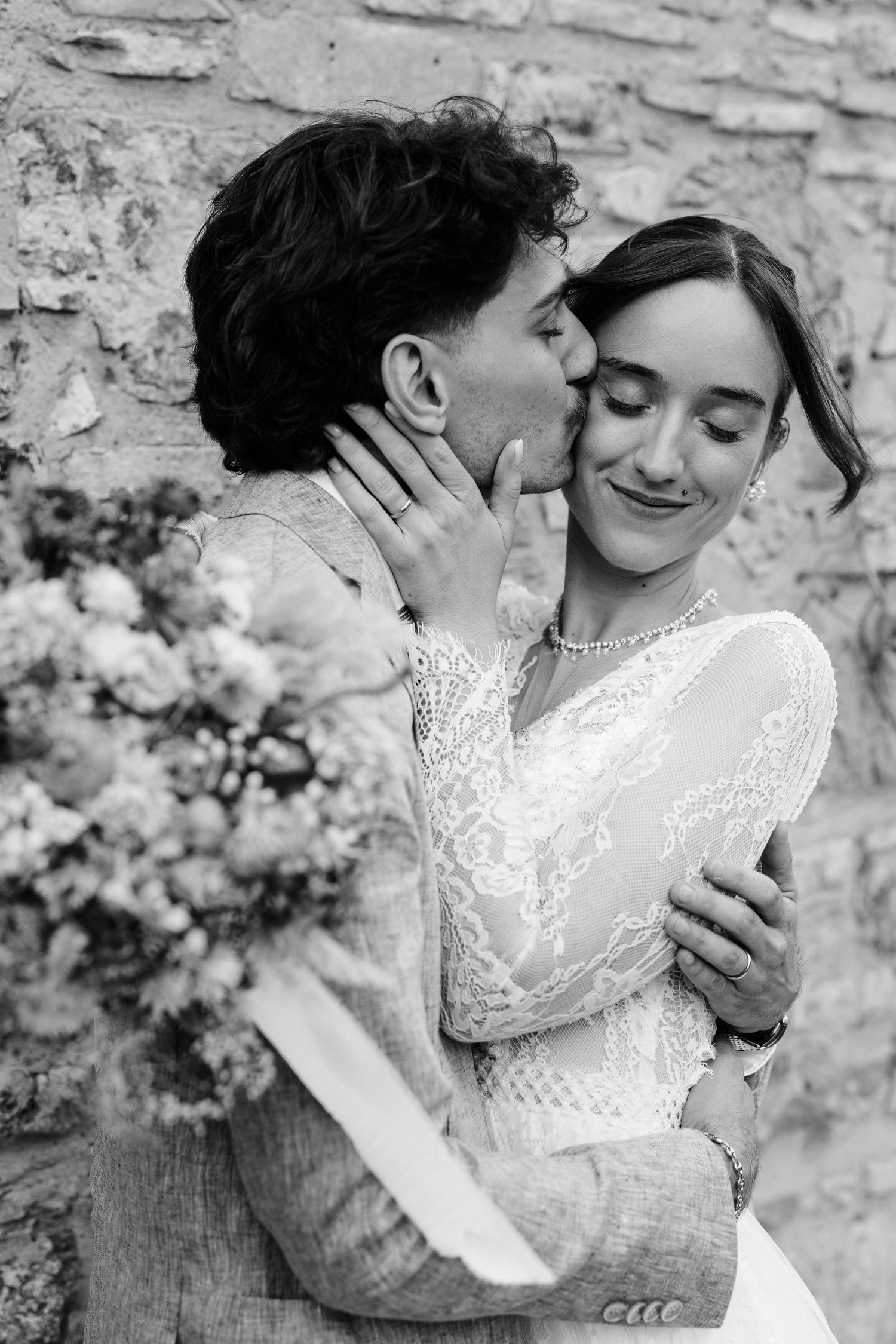 Black and white photo of groom kissing bride on cheek while holding a floral bouquet.