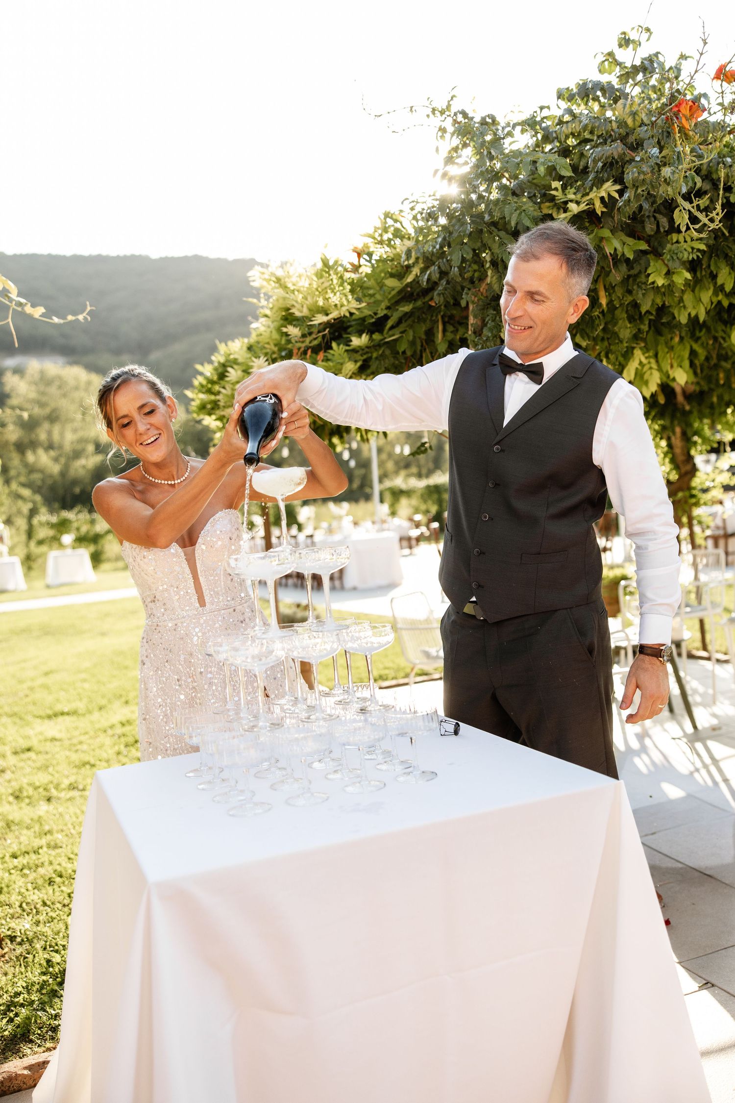 Bride and groom pouring champagne tower at outdoor wedding reception surrounded by lush greenery.
