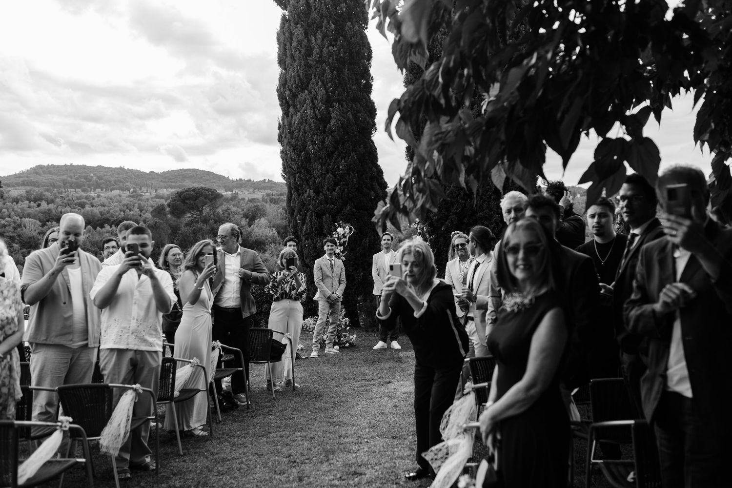 Black and white outdoor wedding ceremony with guests seated and standing among cypress trees in a scenic countryside setting.