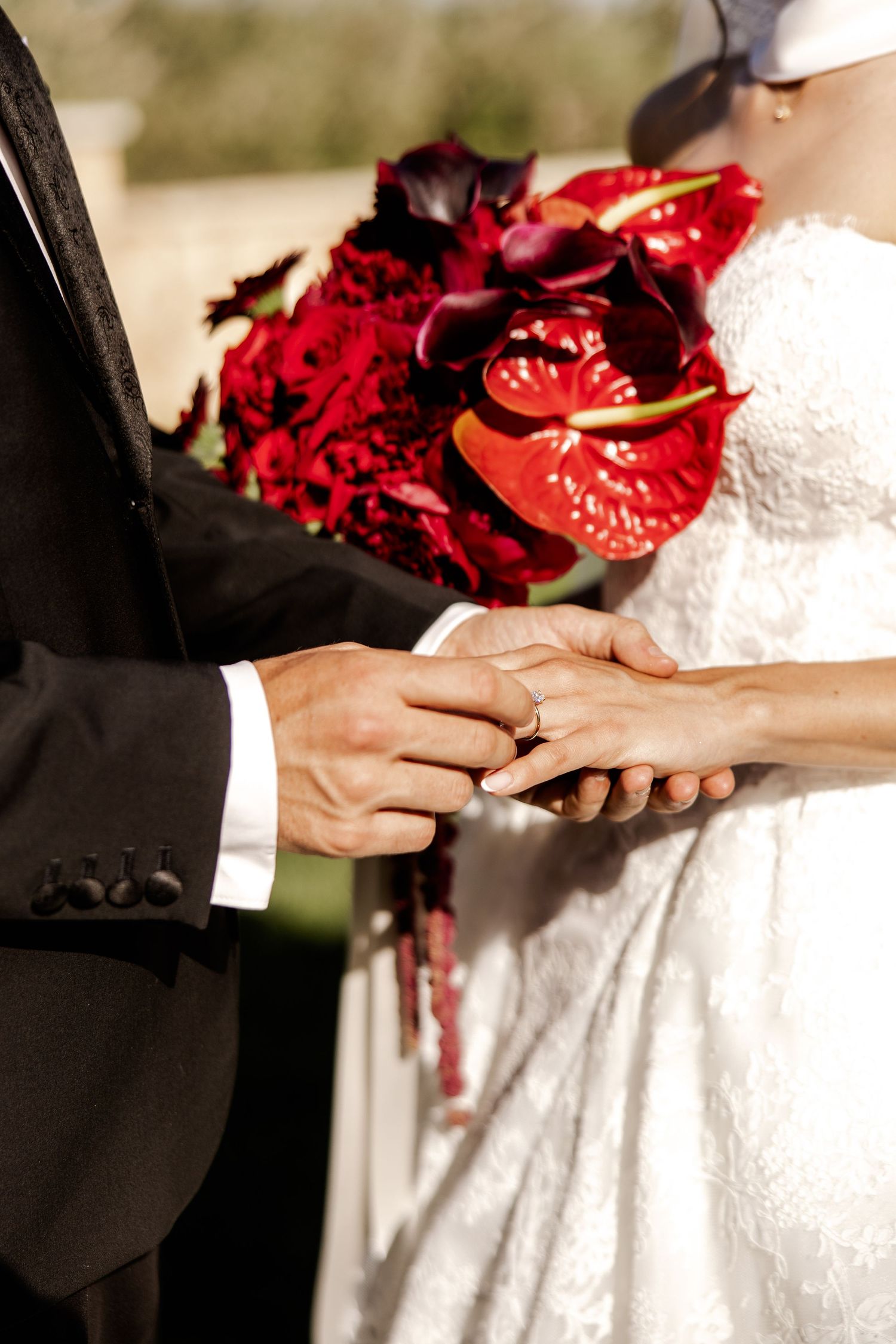 Groom placing a gold wedding ring on brides finger with a vibrant red floral bouquet in the background.
