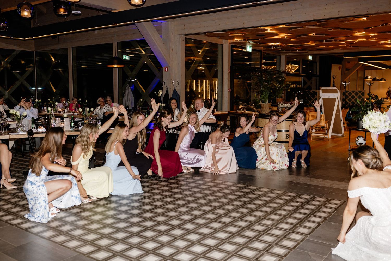 Wedding guests sit on a dance floor with arms raised during a fun bouquet toss reception activity.