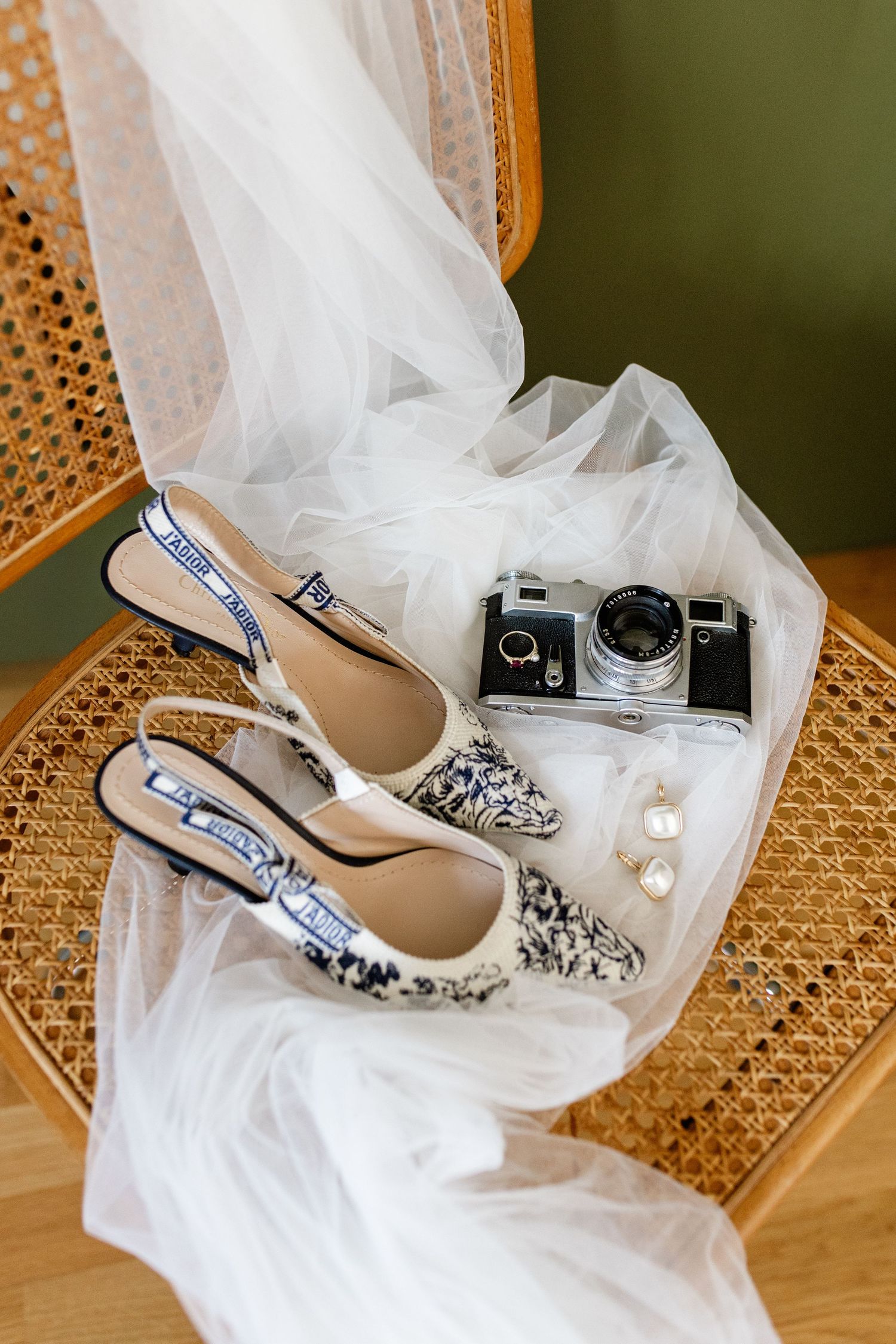 Bridal details flat lay featuring Dior slingback heels, vintage film camera, pearl earrings, and white tulle veil on rattan chair.