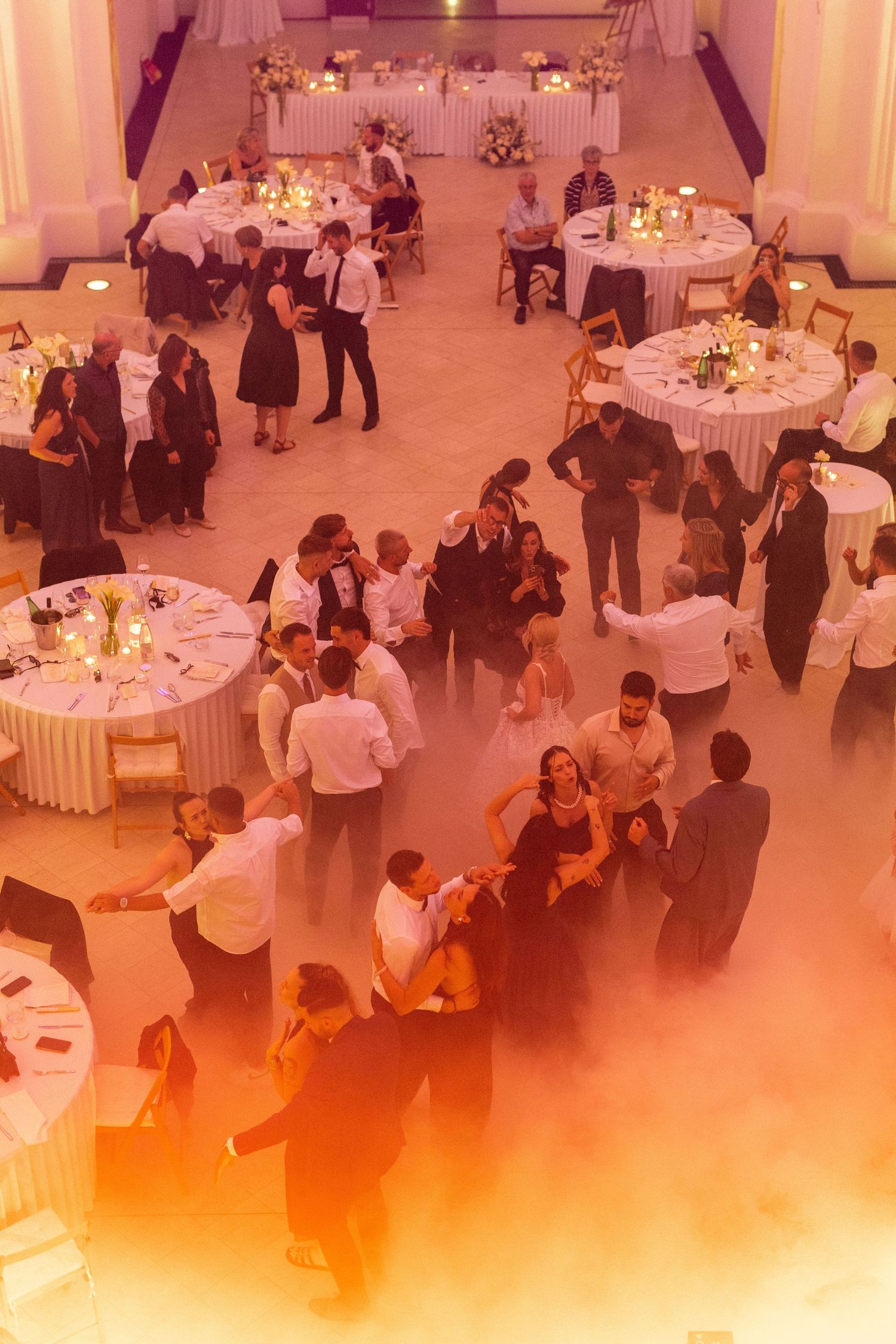 Wedding reception guests dance on a fog-filled dance floor under warm orange lighting in an elegant ballroom.