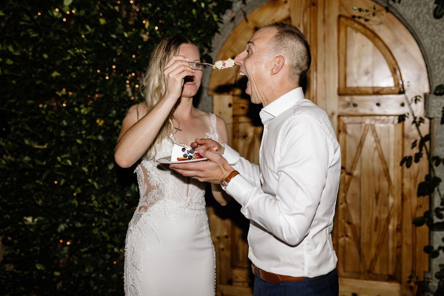 Bride and groom laughing during wedding cake cutting ceremony outdoors at night near wooden doors.