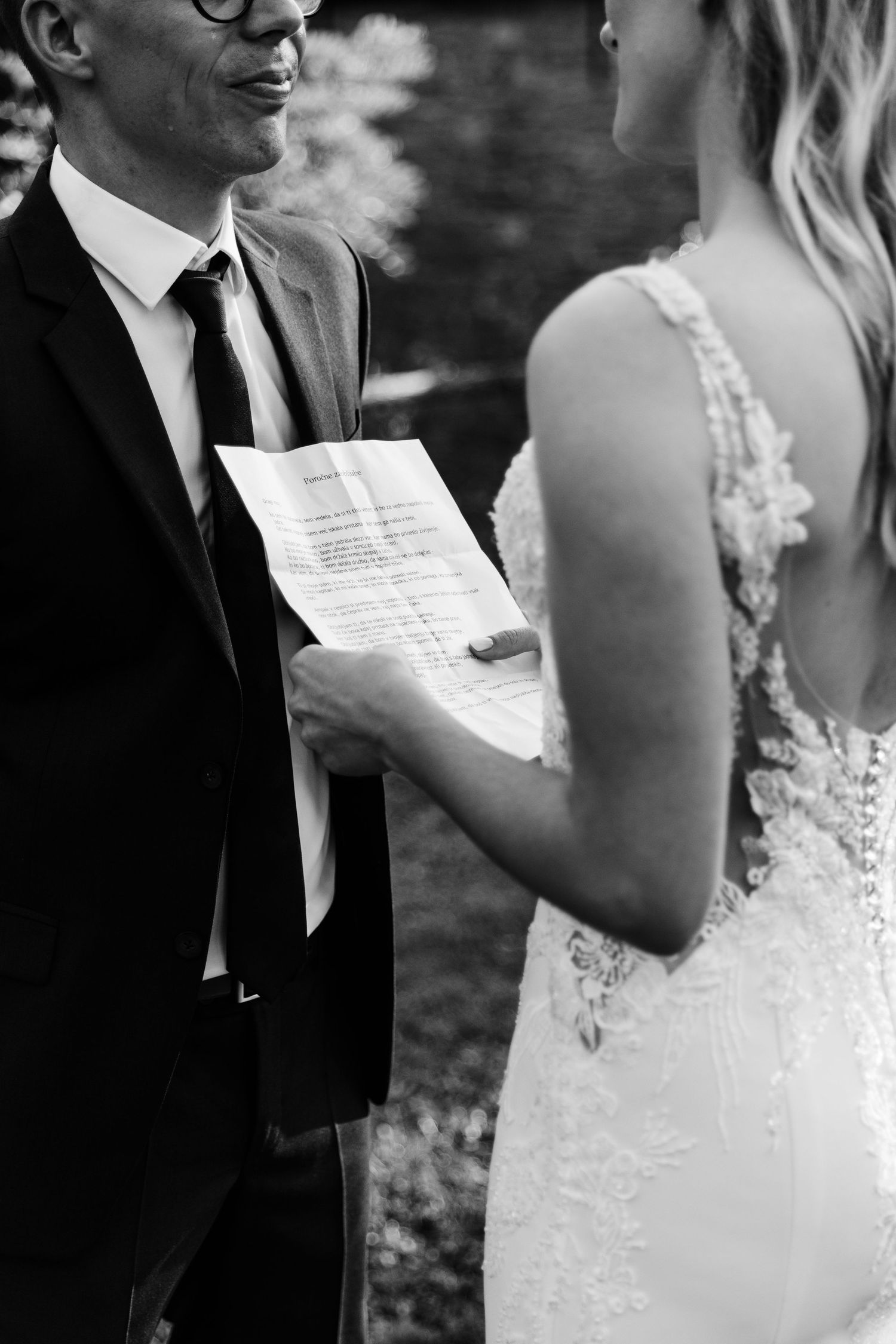 Black and white photo of groom reading wedding vows from paper to bride in lace dress during outdoor ceremony.