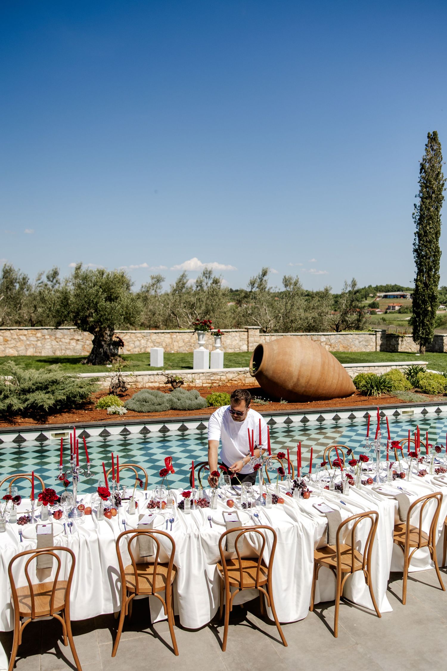 An elegantly set outdoor wedding reception table with red candles and flowers beside a decorative pool in Tuscany.