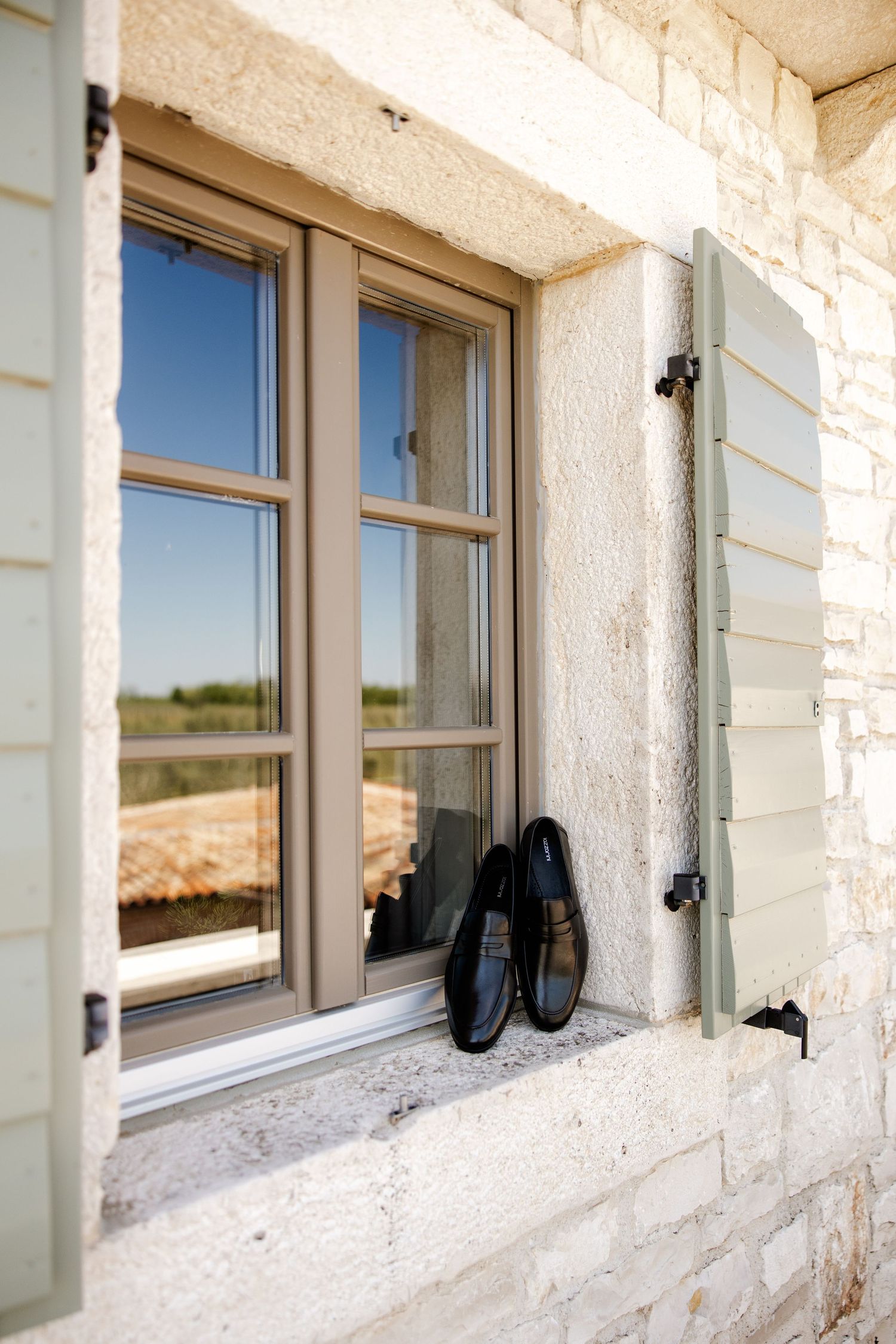 Black leather dress shoes resting on a stone windowsill with green shutters on a rustic exterior wall.